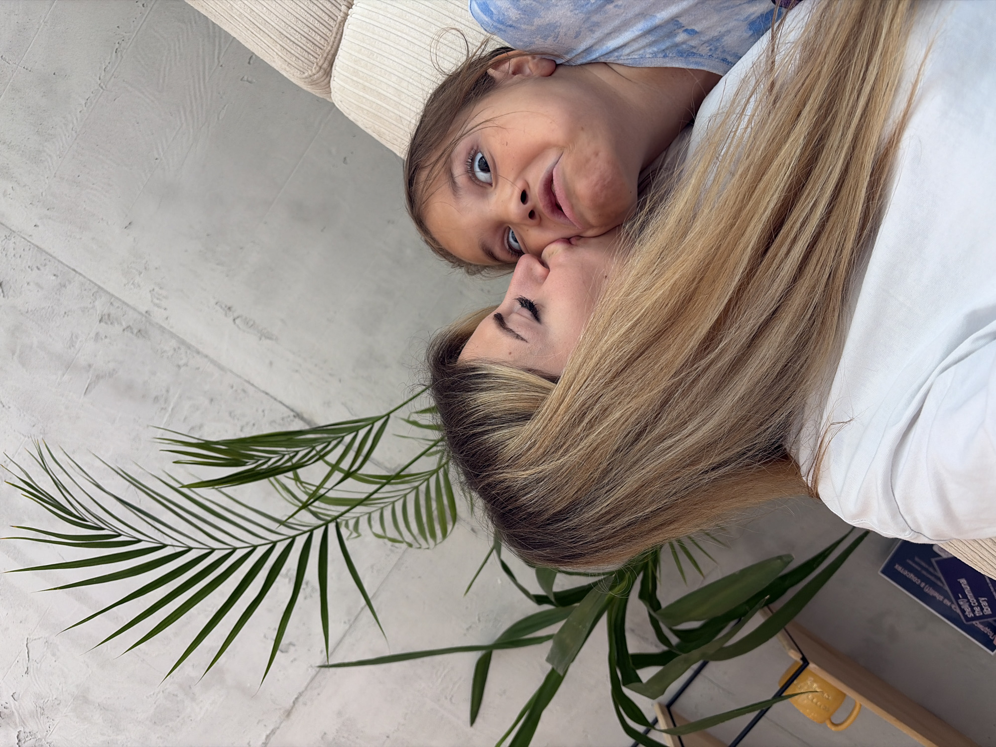 The image shows a close-up of two people, an adult female and a young girl, in what appears to be an indoor setting. The adult female has long, blonde hair and is kissing the girl on the cheek. The young girl has her eyes wide open and a slightly contorted facial expression, as if reacting to the kiss. She is wearing a light blue and white tie-dye t-shirt. The background consists of a textured grey wall, a potted plant with long green leaves, and a portion of a light-colored sofa with a ribbed texture. There is also a wooden shelf with a yellow mug and some books visible. The overall lighting suggests an indoor environment, possibly a living room or a lounge area. The image does not contain any specific visual cues to determine the city of Budva, Montenegro.