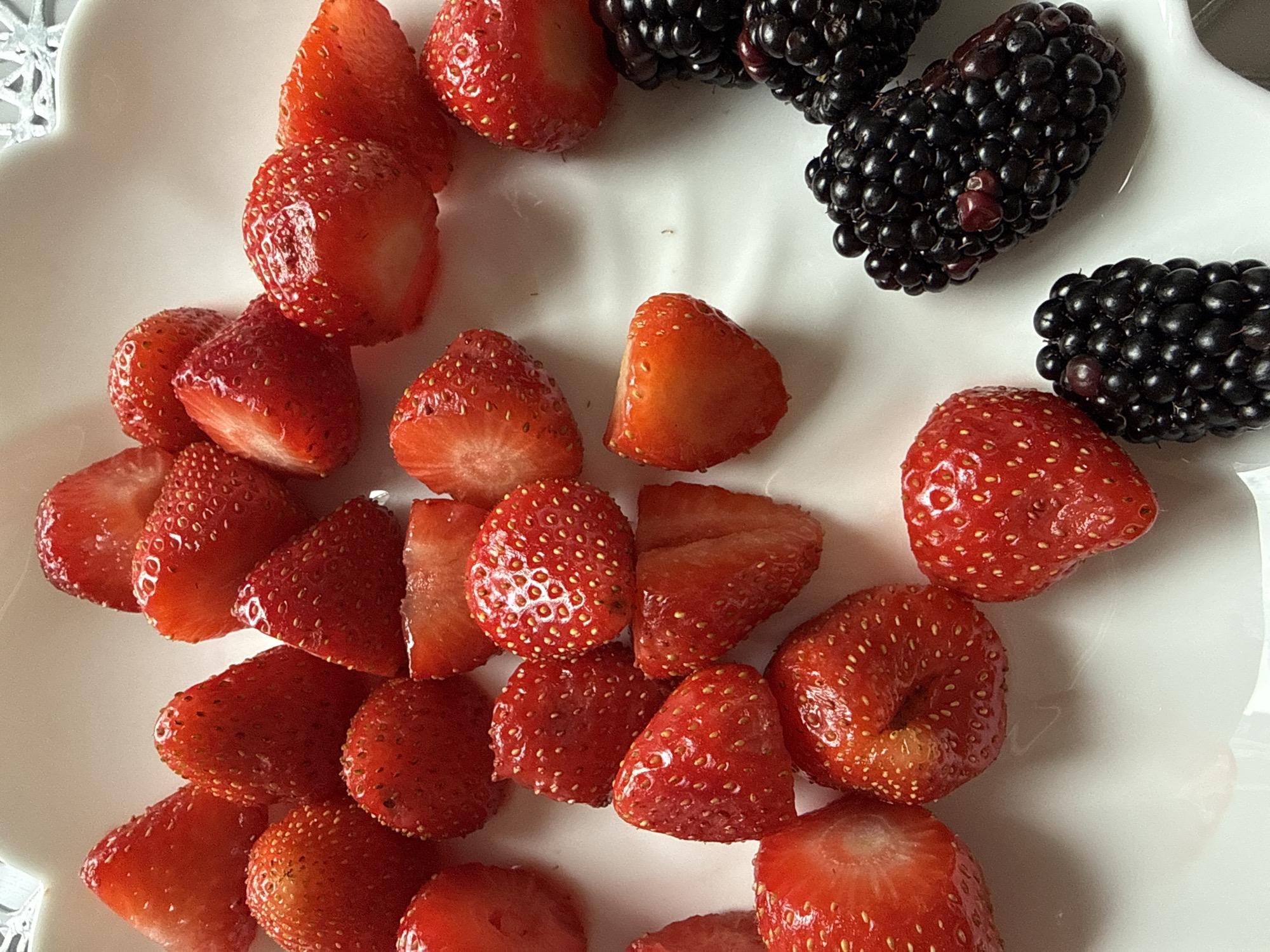 A close-up, overhead shot features a white, scalloped-edged plate laden with fresh strawberries and blackberries. The strawberries are a vibrant red, some whole, others halved, showcasing their seedy texture. The blackberries are a deep, dark purple-black, clustered together. The lighting suggests it is daytime, likely indoors, as there are no discernible outdoor elements to indicate a specific location in Khimki, Russia. The arrangement is simple, focusing solely on the fruit against the plain white of the plate. There are no people or actions depicted.