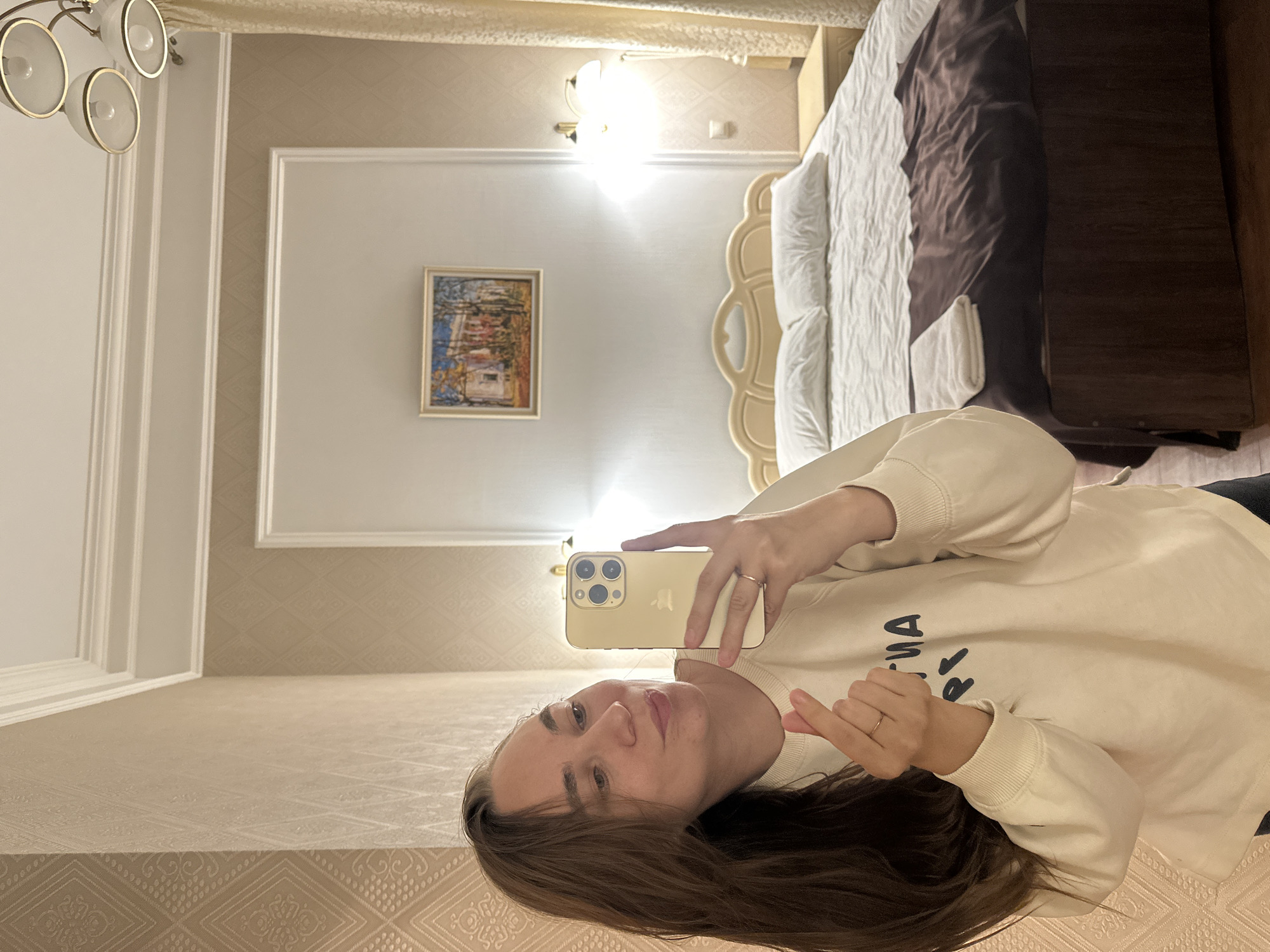 A young Caucasian woman with long brown hair is taking a selfie in a hotel room. She is wearing a cream-colored sweatshirt and appears to be making a heart shape with her fingers. In the background, a bed with white linens and a dark brown throw is visible. A painting hangs on the wall above the bed, and wall sconces provide light. The room has textured wallpaper and decorative moldings. The location is a hotel room in Khvalynsk, Russia.