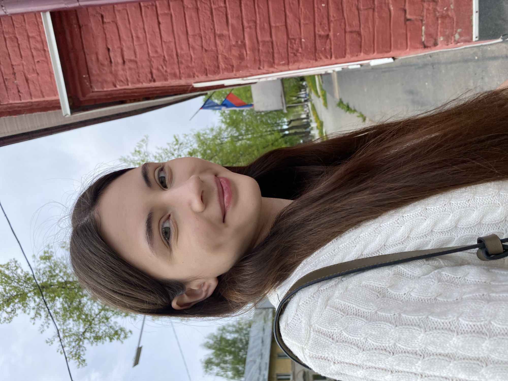 A young, light-skinned woman with long brown hair and fair eyes poses for a photo. She is smiling slightly, wearing a white cable-knit sweater and a shoulder bag with a dark strap.
The background features a red brick building with a white trim. Across the street, a Russian flag is visible near green trees. The scene appears to be Votkinsk, Russia, as indicated by the flag and architectural styles.