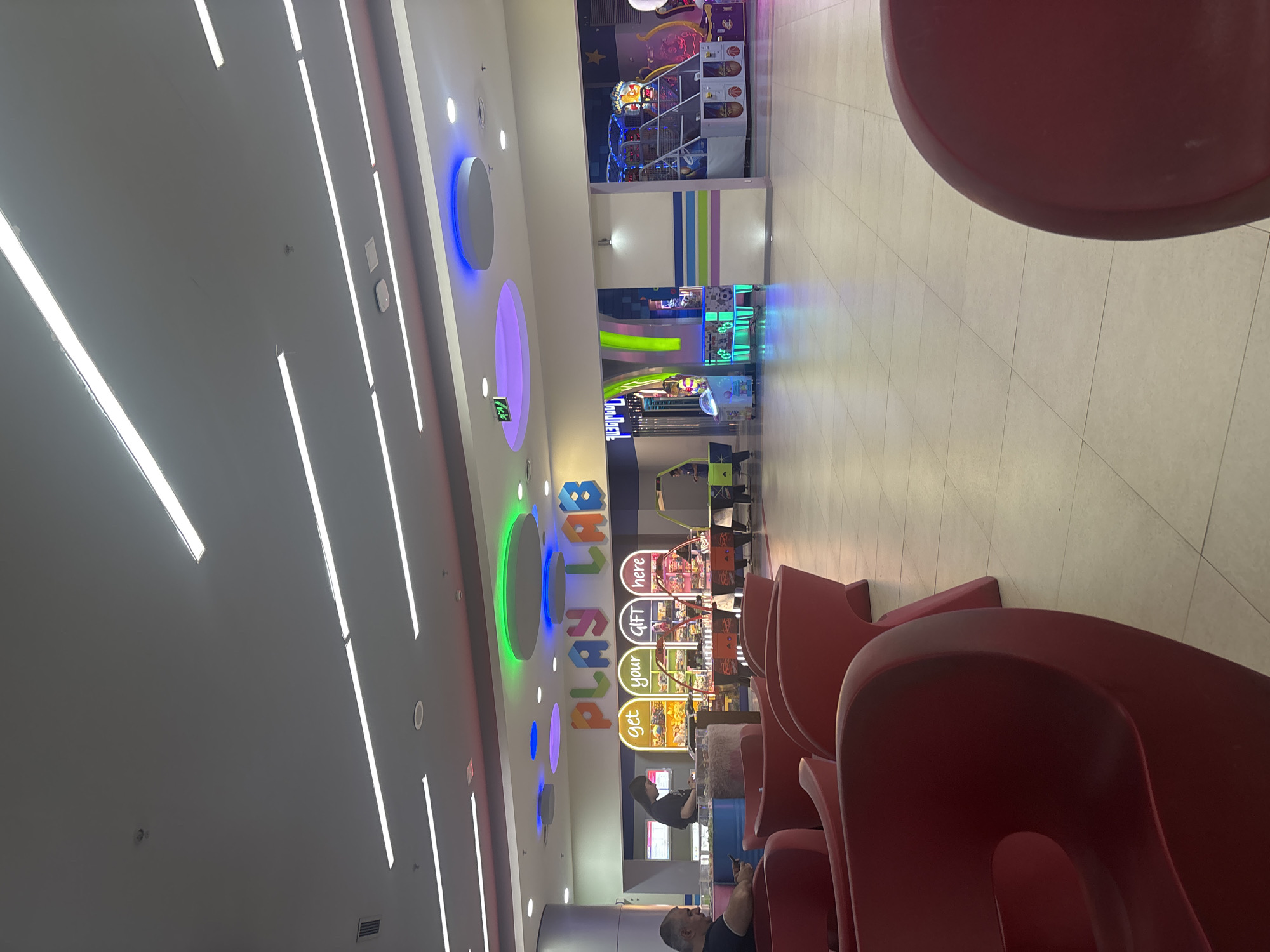 This image captures the interior of a brightly lit entertainment center or arcade, likely situated in Yerevan, Armenia. The scene features a modern design with a light-colored, tiled floor and a grey ceiling. The ceiling is adorned with numerous long, linear white LED strip lights and several large, circular ambient lights casting vibrant blue, purple, and green glows.
In the foreground and midground, a series of glossy, ergonomically shaped red chairs or seating units are arranged. One person, appearing to be an adult male, is seated in a red chair in the immediate foreground, looking down at a mobile device. Another individual is faintly visible further back, near the gaming area.
The background is dominated by various arcade machines, including a basketball-themed game and other colorful redemption games. A prominent, multi-colored sign on the back wall spells out "PLAY LAB." Below this main sign, additional smaller signs read "get," "your," "GIFT," and "here," indicating a prize counter or reward system. The overall ambiance suggests a lively yet comfortable leisure environment, with people relaxing and engaging with the available entertainment. The time of day is indeterminate due to the indoor setting with artificial lighting.