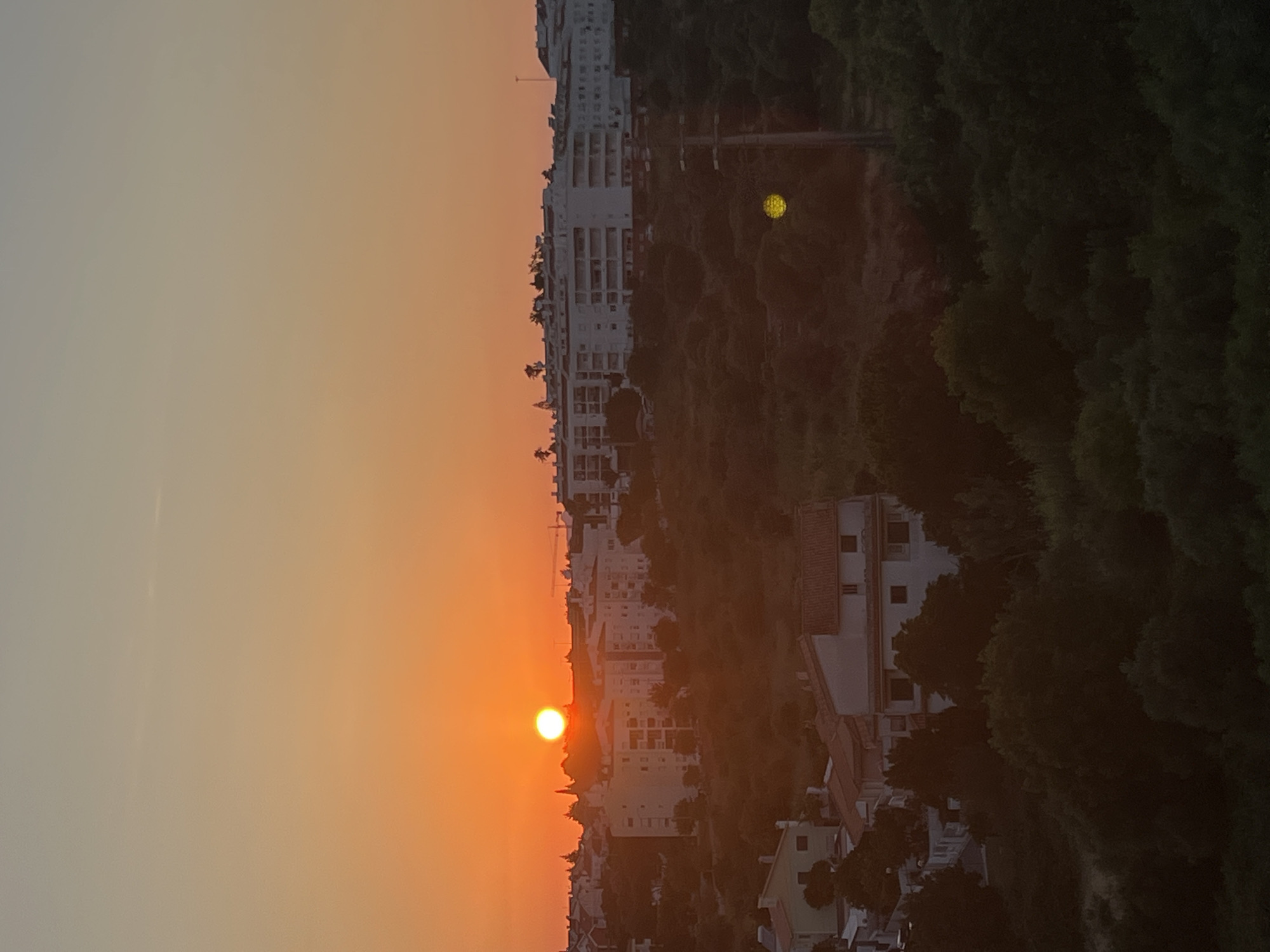 An outdoor, medium-angle, vertical shot captures a sunset over a residential area in Cascais, Portugal. The sun, a bright orange orb, is low on the horizon, casting a warm glow across the sky, which transitions from orange near the sun to a pale grey at the top. A cluster of white, multi-story apartment buildings dominates the mid-ground, with smaller residential houses visible in the foreground and on the left side of the frame. These structures are nestled among lush, dark green trees that cover the rolling hills. A utility pole with crossbars is visible on the right side of the image, and a bright yellow circular light reflection is present in the foreground. No people are visible in the image.