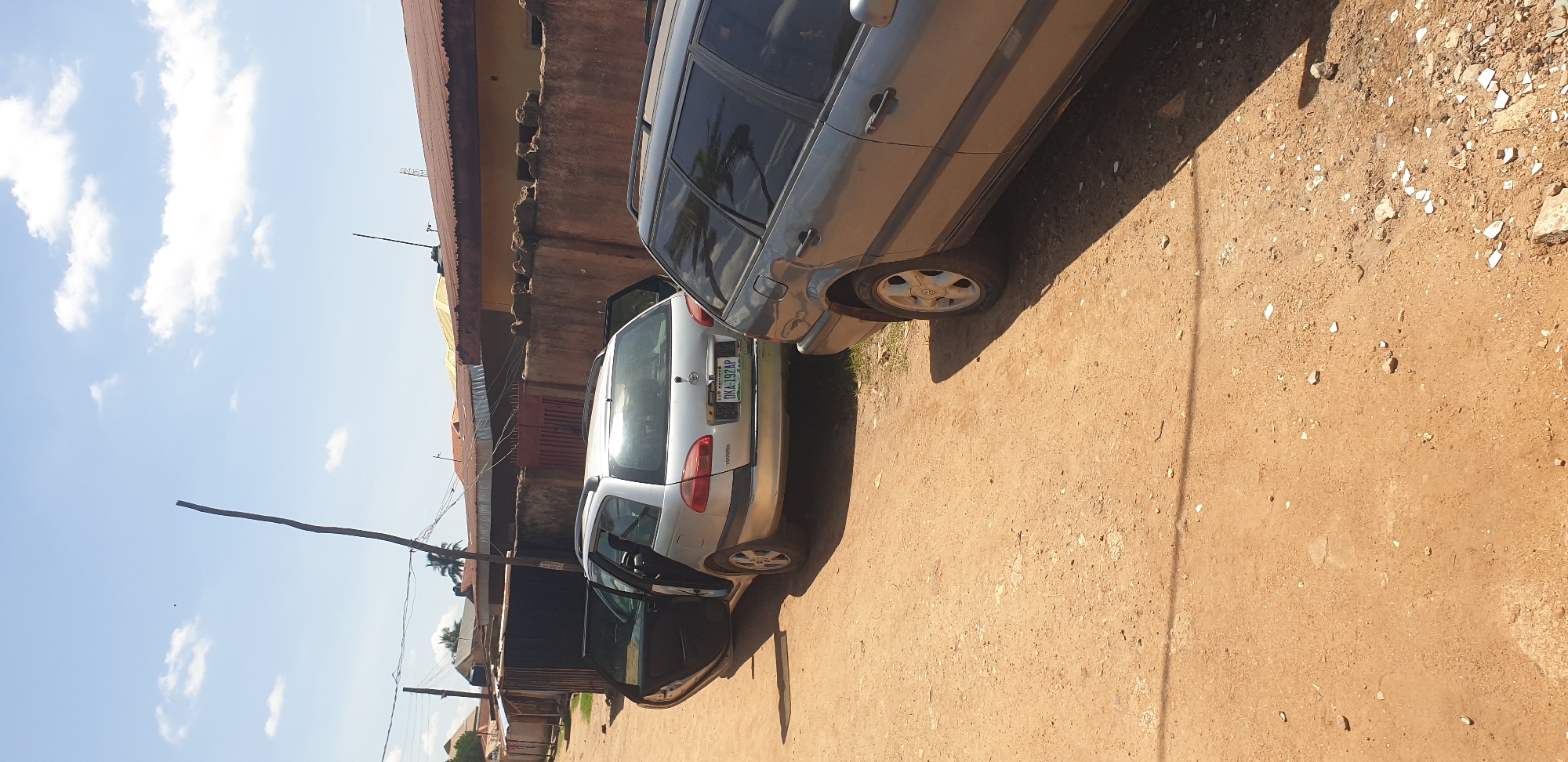 The image depicts an outdoor scene in Sabon Tasha, Nigeria, under bright, clear conditions, likely midday or early afternoon, as evidenced by strong shadows. The setting is an unpaved, reddish-brown dirt path or road. Two station wagon vehicles are parked on the path.
The primary subject is a silver Toyota station wagon (identified by the "Toyota" emblem and license plate details) positioned in the center-left. Both its front and rear passenger-side doors are open. The vehicle displays a Nigerian license plate, "DKA-192AP," with "KADUNA" visible below, indicating registration in Kaduna State. To the right, partially visible in the foreground, is a dark grey station wagon, appearing to be a Volkswagen Passat variant, with its doors closed. Both vehicles are equipped with roof rails.
The background features a low, reddish-brown concrete or mud wall with an uneven top, bordering a property. Behind this wall and further in the distance are several buildings with corrugated metal roofs, some rusted brown and others a faded yellow. A dark metal gate is partially visible behind the silver car, leading into a property. A tall, slender wooden pole, appearing to be a stripped tree trunk, stands prominently on the left side of the scene, extending upwards into the bright blue sky, which has scattered white cumulus clouds. Utility wires are strung across the upper portion of the image. Sparse vegetation, including some grass and a distant palm tree, is visible. No individuals are present in the scene.