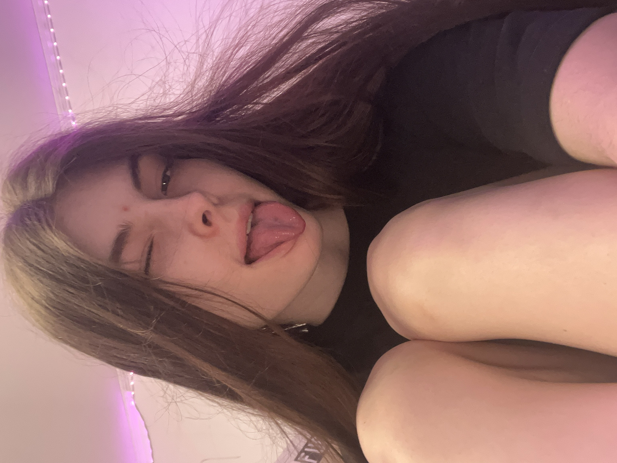 A young woman with long brown hair is lying down, looking up at the camera. She is sticking out her tongue and winking with her left eye. She is wearing a black shirt and has fair skin. The background appears to be a bedroom setting, with a string of lights visible along the top edge of the frame. The lighting is soft and pinkish. There is no evidence to determine the location in the city of Saint Petersburg, Russia.