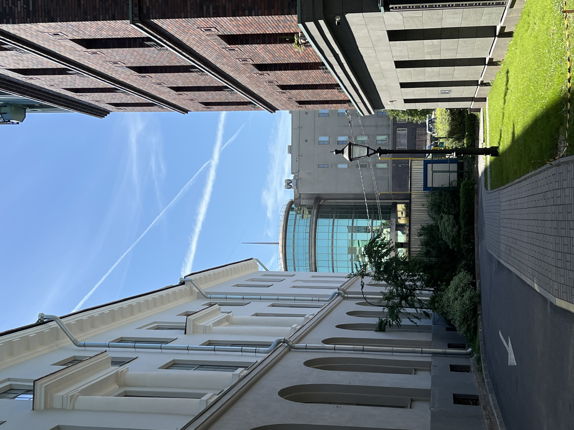 The image depicts an urban scene in Saint Petersburg, Russia, likely on a bright, sunny day given the clear blue sky and strong shadows. The perspective is from a pedestrian walkway, looking down a path bordered by buildings and greenery.
On the left, a white building with classical architectural elements like columns and arched ground-level windows is visible. This building has a light grey base and a white upper section with a decorative cornice. Rain gutters are prominently featured along its facade.
To the right, a dark brick building with a more modern, severe design stands tall. It has a pattern of small, square openings or decorative elements interspersed with the brickwork. The facade also features a dark grey stone base.
In the mid-ground, a distinct circular building with a glass facade, reflecting the blue sky, is prominent. It appears to be a modern structure. Behind it, a grey, multi-story building is visible.
A black lamppost with a classic design stands along the pathway. The path itself is paved with light-colored rectangular bricks, leading into the distance. To the right of the path, a patch of well-maintained green grass is illuminated by sunlight, casting a distinct shadow.
In the distance, some trees and other foliage suggest landscaping or a park area. There are also hints of vehicular traffic and possibly other urban infrastructure. The sky above is a clear blue with prominent white contrails from aircraft, forming an X-shaped pattern.
There are no people or animals visible in the frame. The scene conveys a sense of an architectural ensemble in a developed urban environment.