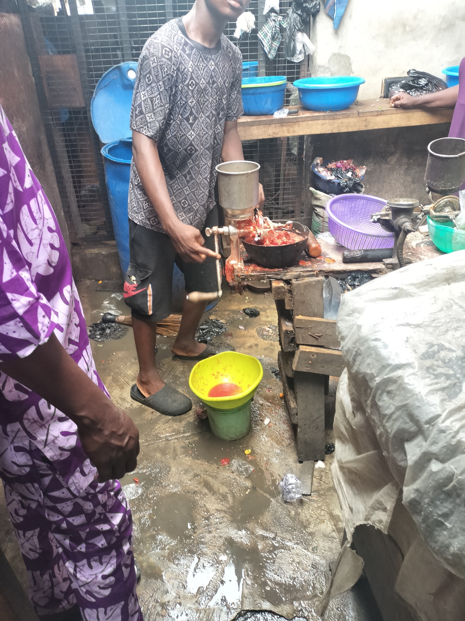 The image depicts a wet, possibly outdoor food preparation area in what is likely Alaiyabiagba, Nigeria. A young man wearing a patterned shirt and shorts operates a manual food grinder, processing a red, pulpy substance (likely tomatoes or peppers) into a bowl. A woman stands nearby, wearing a purple and white patterned garment. There are blue tubs and various bowls on a rough wooden table in the background. The ground is wet and littered with food debris. The environment appears to be a busy commercial space or market.