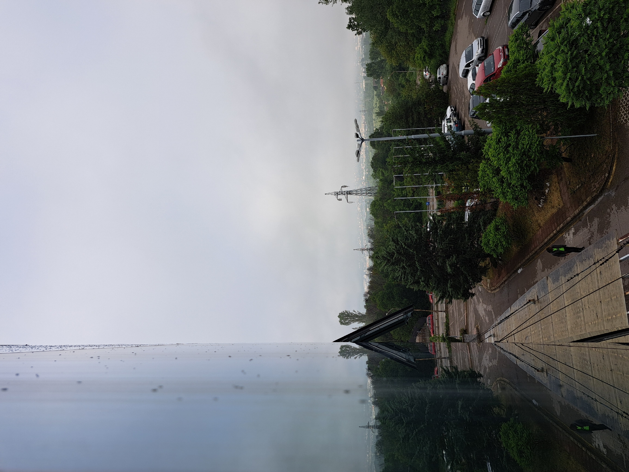 This image captures an outdoor urban or suburban landscape from an elevated perspective, likely a building window or balcony. The foreground is dominated by a reflective glass surface, possibly a window, which is covered in numerous small water droplets, indicating recent rain. This surface reflects a distorted view of the sky and the scene below.
Beyond the reflective surface, the scene unfolds with a paved area or road on the right, leading into a parking lot filled with parked cars of various colors, including white, silver, red, and dark hues. Dense green trees line the road and dot the landscape, some appearing to be conifers. Two individuals are visible; one stands on the paved area near the building, wearing a dark jacket and a high-visibility green or yellow vest. Another person, also in a reflective vest, is partially visible near cars further away.
In the mid-ground, a tall lamppost with multiple lights stands prominently. Further in the background, several large power line pylons rise above the tree line. The sky is uniformly overcast and bright, suggesting daytime but with heavy cloud cover. The wet appearance of the roads and paved surfaces, along with the water droplets on the glass, confirms recent rainfall. The distant horizon reveals a sprawling city or town landscape under the cloudy sky. No discernible text is visible in the image. The location context provided indicates the scene is in Sofia, Bulgaria.
