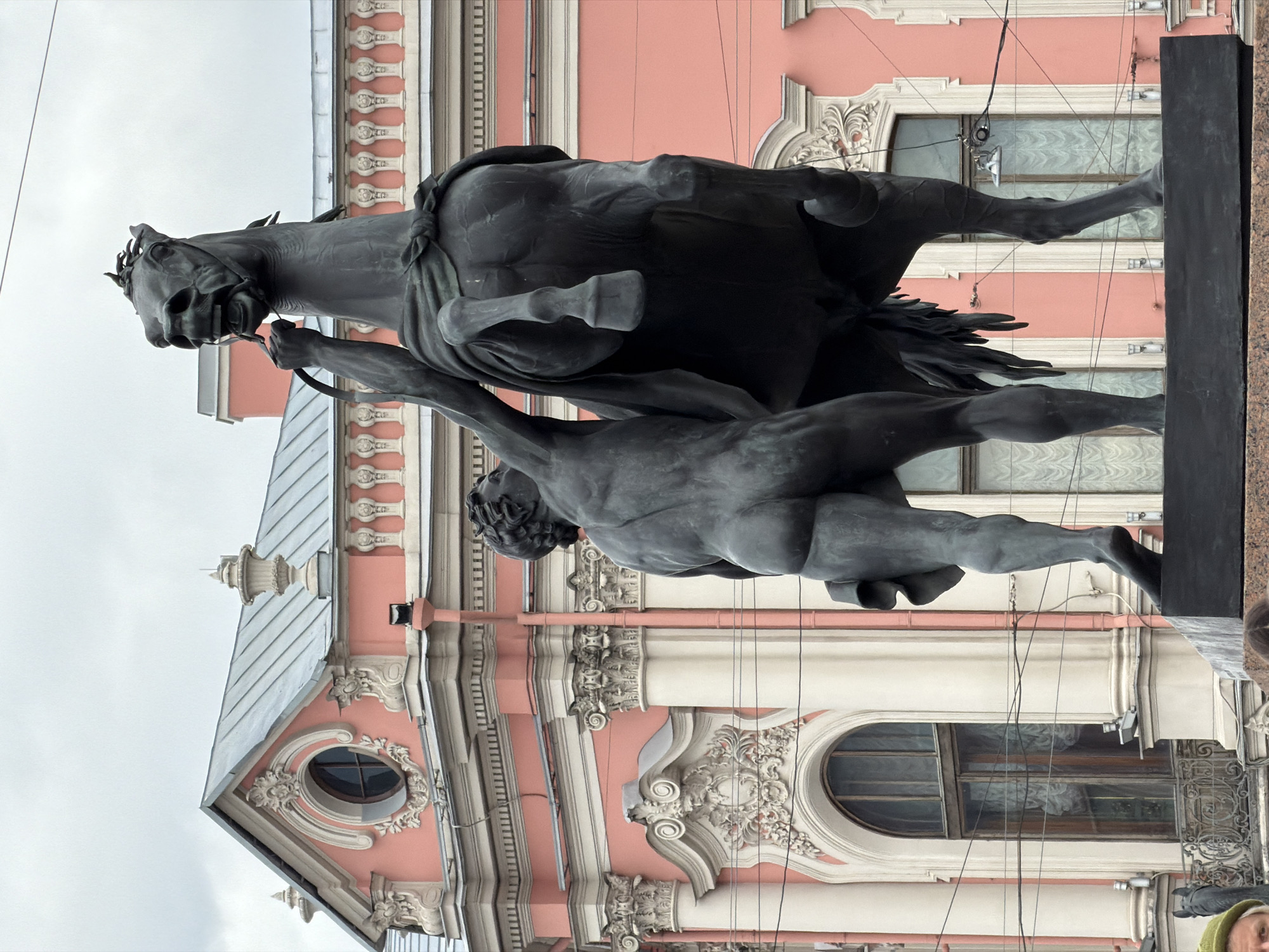 The image displays a dramatic bronze equestrian statue set against the backdrop of an ornate, light pink historical building in Saint Petersburg, Russia. The central subject is a powerful, dark-colored horse rearing up on its hind legs, with its head turned sharply to the left, mouth agape, and an expression of struggle. A muscular, nude human figure is depicted alongside the horse, attempting to control it with reins or straps. The human figure's pose suggests a dynamic struggle, with taut muscles and a determined effort to restrain the powerful animal.
The background building is a striking example of classical architecture, featuring a pale pink facade adorned with intricate white stucco decorations, including pilasters, Corinthian columns, ornate window frames, and decorative moldings. Several windows, both arched and oval-shaped, are visible, reflecting the cloudy sky. The building's roofline is characterized by a grey, pitched roof and decorative finials.
The sky is overcast, indicating a cloudy day. Overhead wires, possibly for trams or trolleybuses, crisscross the scene. In the bottom right corner of the main image and one crop, a portion of a person's head wearing a green hat is faintly visible, suggesting human presence near the monument. No discernible text is visible on the statue or the building. The scene captures a moment of intense action frozen in bronze, contrasting with the static, elegant architecture behind it.