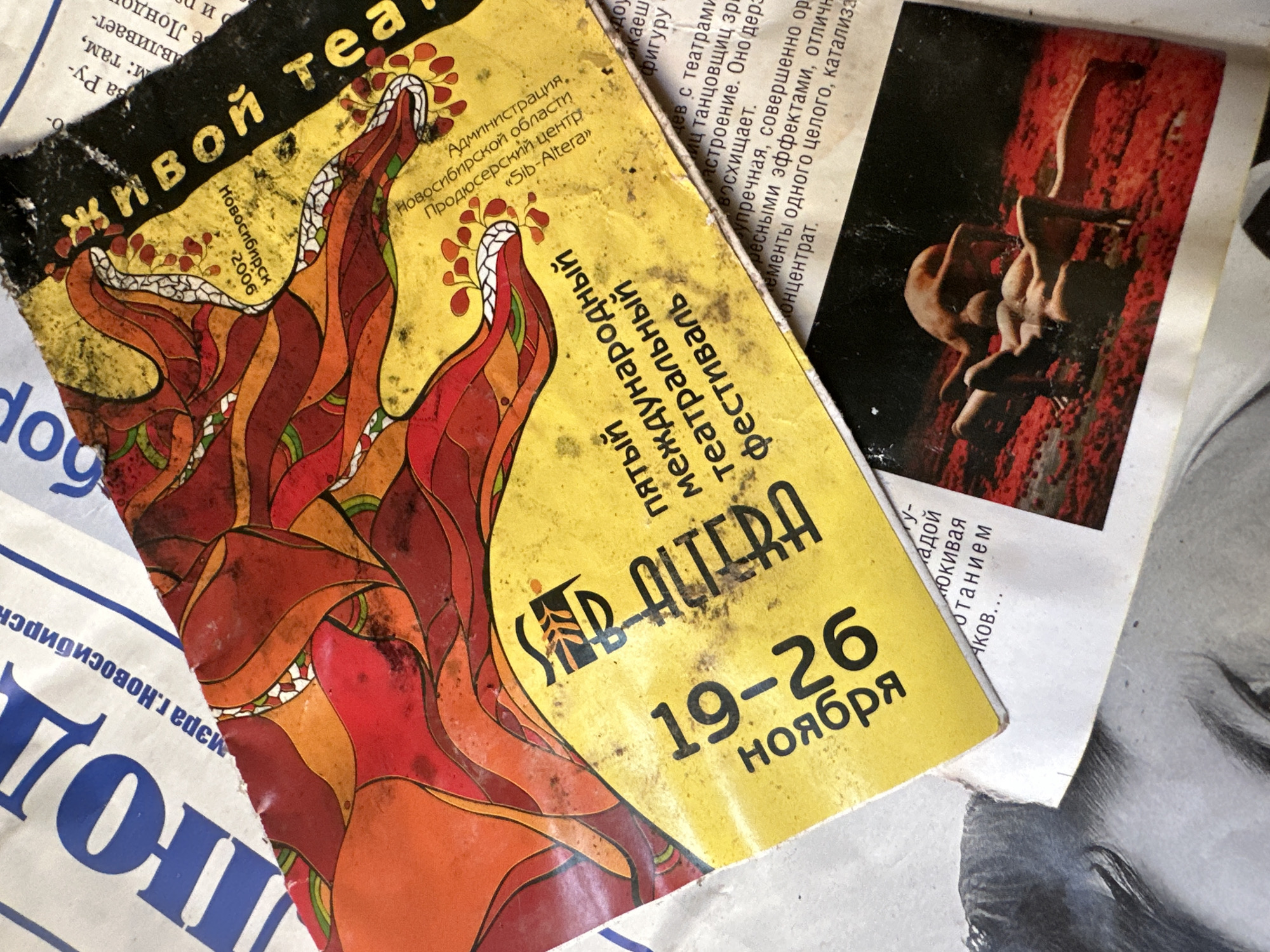 This image shows a close-up, overhead view of a yellow flyer or poster, partially unfolded and resting on top of several newspapers. The flyer features a vibrant, abstract illustration of intertwined red, orange, and yellow organic shapes, possibly representing flames or stylized serpents, against a speckled yellow background. In large, stylized black lettering, the text "АРТ ДИТРА" is visible. Below this, "ПЯТЫЙ МЕЖДУНАРОДНЫЙ ТЕАТРАЛЬНЫЙ ФЕСТИВАЛЬ" is written in smaller black text. The dates "19-26 НОЯБРЯ" are prominently displayed in large black numerals and text on the left side.
Further text in smaller black lettering is scattered across the flyer. One section reads "Администрация. Новосибирской области." and "Продюсерский центр. «Sib-Altera»". Another section, partially obscured, appears to contain descriptive text about theatre. In the upper right corner, partially visible black text reads "живой теа".
The flyer is creased and shows signs of wear, with some discoloration and scuff marks. It's positioned over a newspaper with blue lettering that says "Люди" and "изра г.Новосибирск". A small, rectangular photograph is also partially visible beneath the flyer, showing two nude figures in a pose on a red, textured surface. The overall scene suggests a collection of printed materials, possibly discarded or stored together. The image does not provide any specific information about the city of Biysk, Russia; it only contains references to Novosibirsk.
