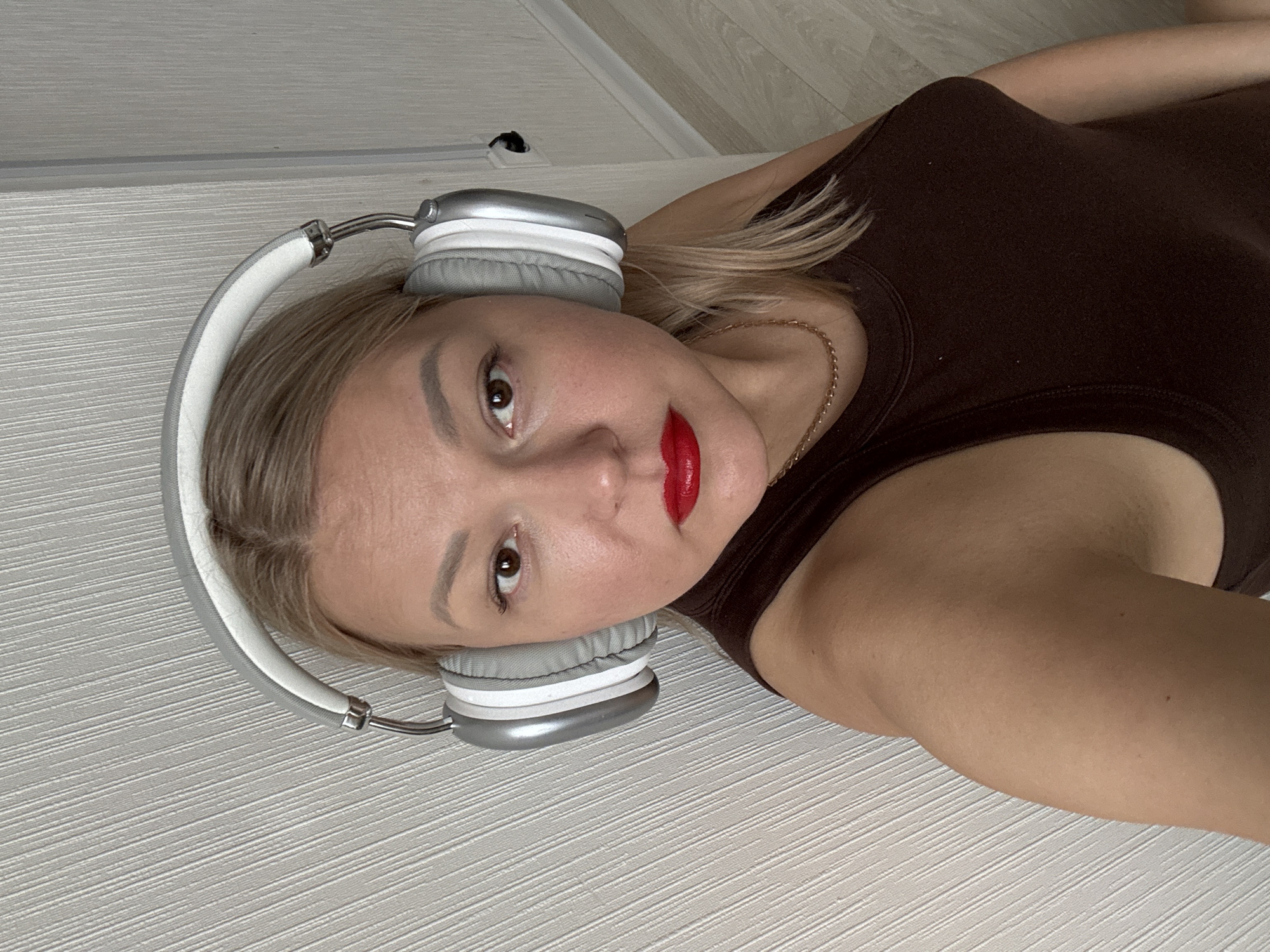A woman is lying on a textured, light-colored surface, possibly a floor or bed. She is wearing headphones with silver and white accents, a dark brown top, and a gold chain necklace. Her hair is blonde, and she has on red lipstick. The background includes a light-colored wall and a power outlet. The image does not contain enough information to determine the location in Izhevsk, Russia.