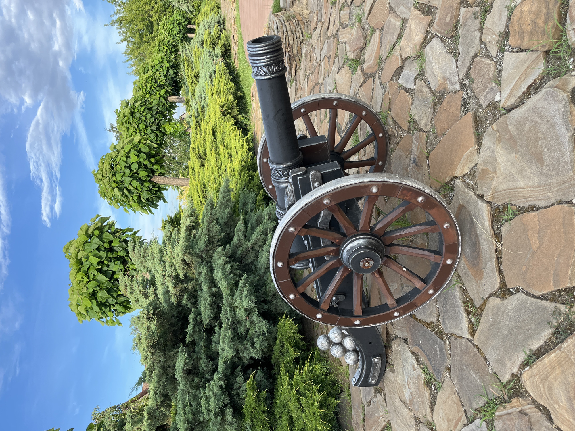 The image depicts a cannon, likely a replica or decorative piece, positioned on a stone pathway. The cannon is black with wooden wheels, featuring decorative metal accents. Beside the cannon, there are several cannonballs arranged in a holder. The setting appears to be an outdoor area with lush greenery, including trees and bushes, suggesting a park or garden. The sky above is blue with scattered white clouds, indicating fair weather. The orientation of the image is vertical, with the cannon situated in the lower right quadrant. The stone pathway is made of irregular, flat stones in earthy tones. The time of day appears to be daytime. Visible text includes "LOUIS XIV" on the cannonball holder. The location context is Azov, Russia.