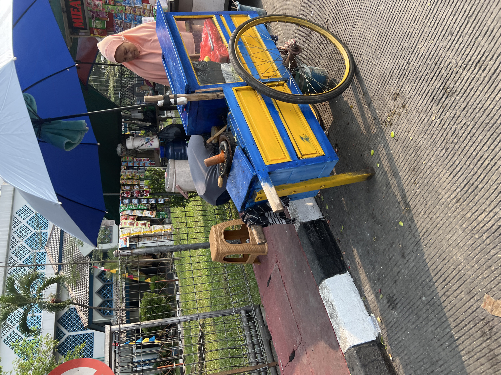 In Citeureup, Indonesia, a street vendor stands beside a vibrant blue and yellow cart under a large umbrella. The cart displays various goods for sale, with a woman in a peach-colored hijab attending to the stall. The background reveals a building with intricate white and blue patterned windows, accompanied by lush green grass and trees, suggesting an outdoor setting in a semi-urban or commercial area. A metal fence separates the street from the grassy area. The road in the foreground is paved with textured asphalt and features a distinctive black, white, and red curb. Scattered leaves and debris are visible on the ground.