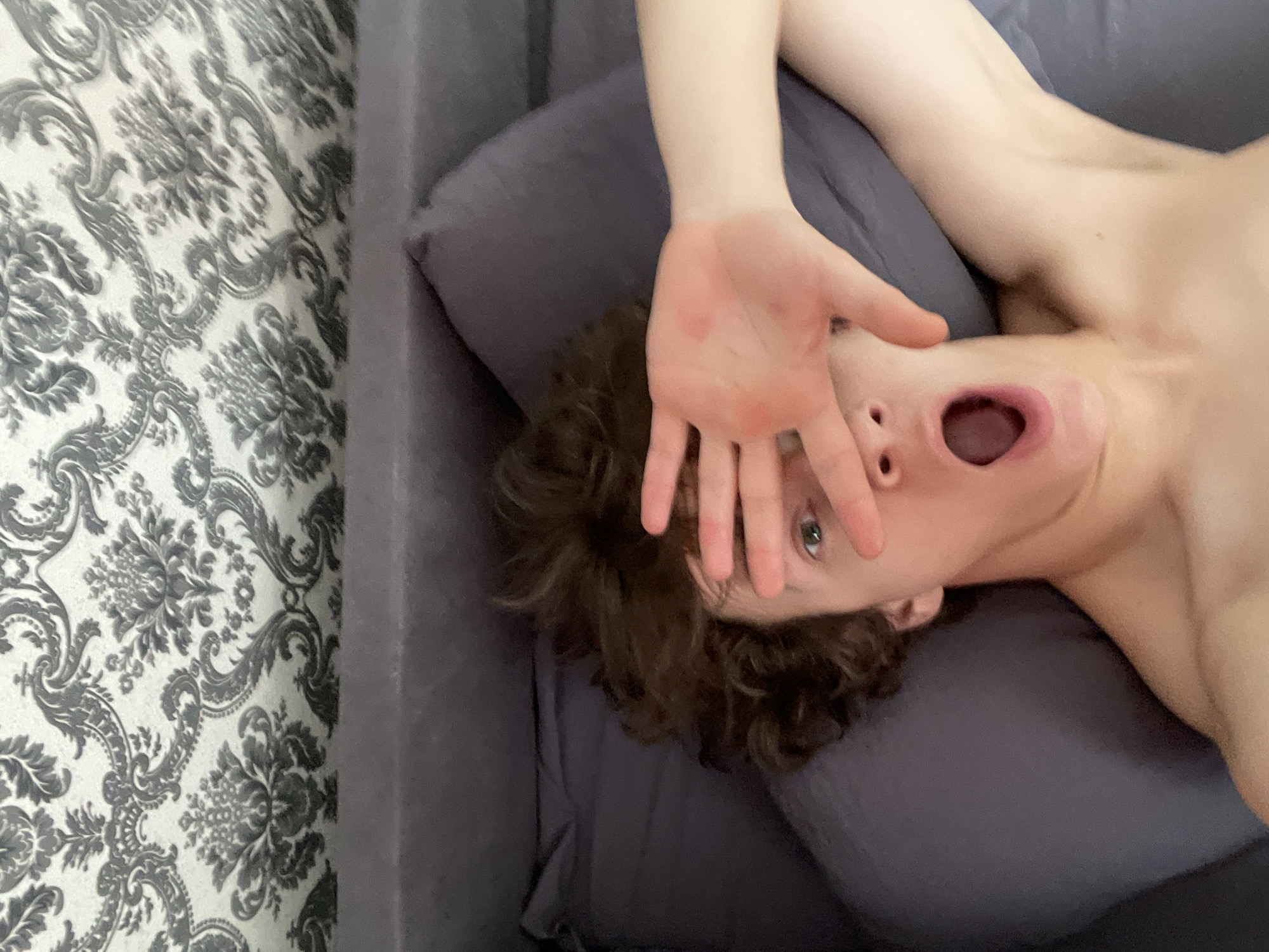 A shirtless, light-skinned person with brown curly hair is lying upside down on a purple couch in an interior space. The person's right hand is raised to partially obscure their face. The person has an open mouth, and their right eye is visible between their fingers. To the left of the couch is a wall covered with a patterned wallpaper featuring floral designs in shades of gray and white. No specific information suggests the location is Krasnoyarsk, Russia.