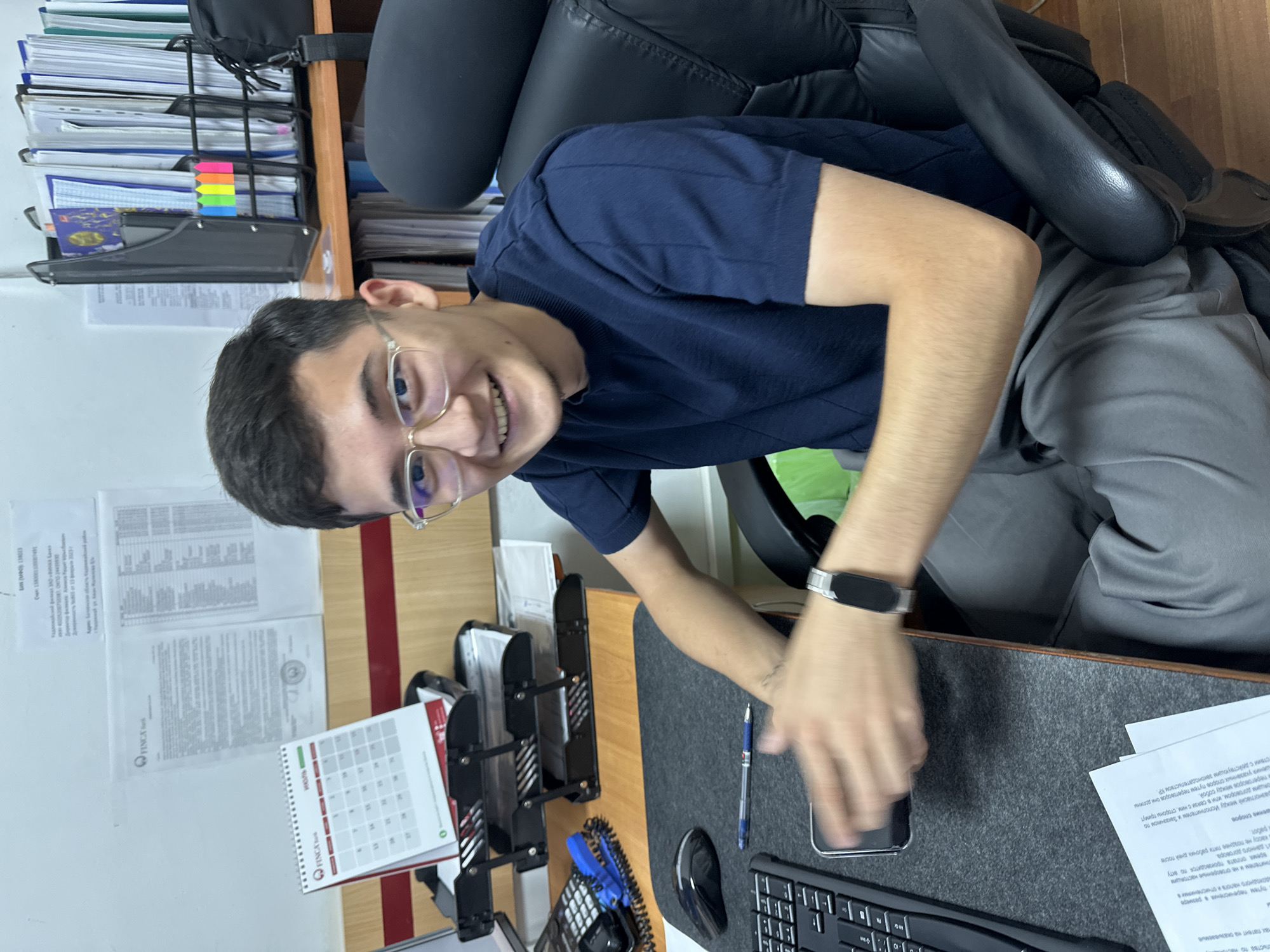 A young man with glasses is sitting at a desk, smiling at the camera. He is wearing a dark blue t-shirt and gray pants. The desk is cluttered with office supplies, including a keyboard, a mouse, a phone, and a calendar. In the background, there are shelves filled with binders and folders. The scene appears to be an office in Kadamjay, Kyrgyzstan.