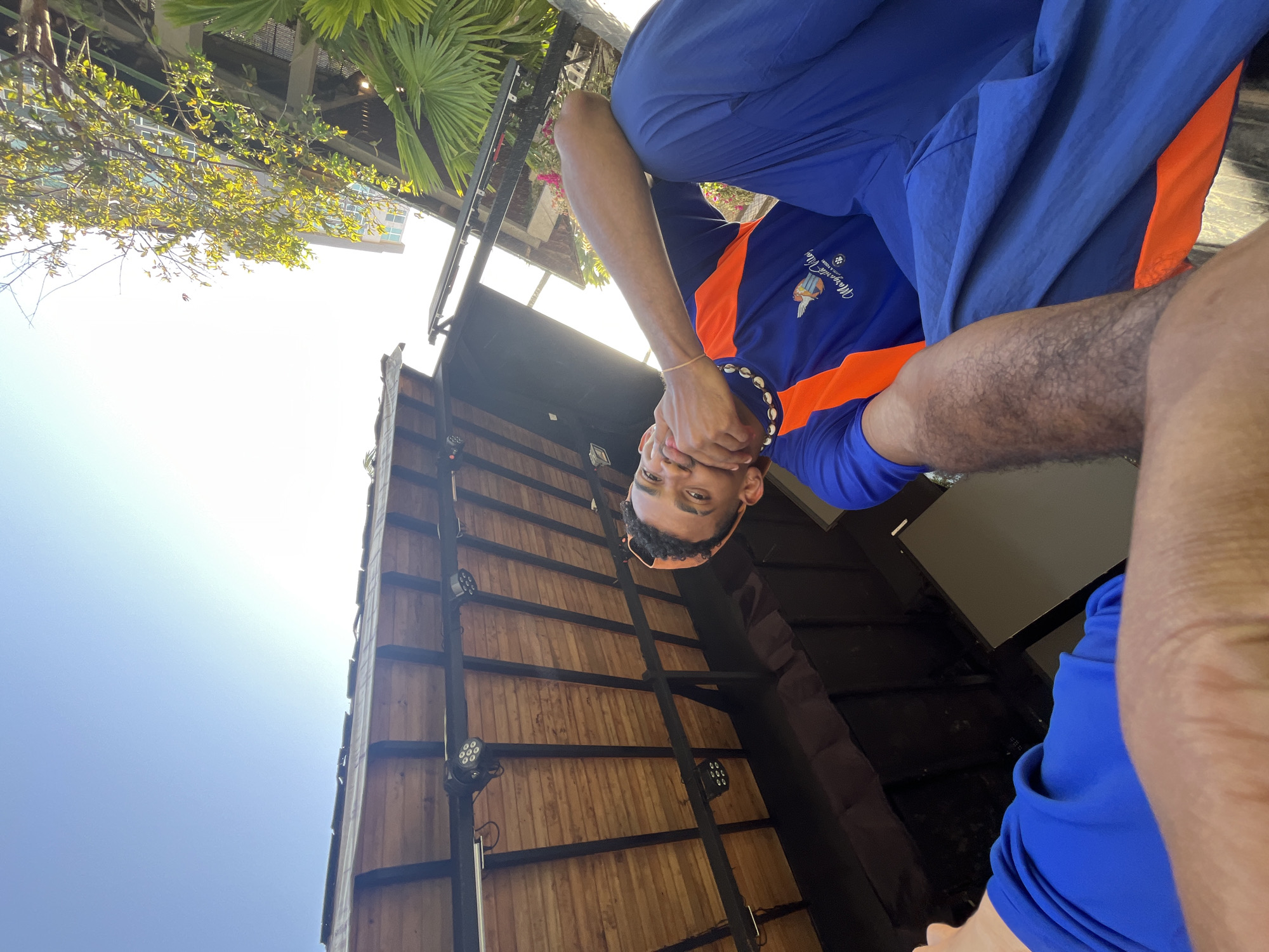 A man wearing a blue and orange shirt with "Margaritaville" printed on it is in the foreground. He's holding his hands in front of his face, wearing what appears to be a beaded headband or necklace. Behind him is a tall, narrow structure with a wood panel facade and dark metal supports. Mounted on the metal supports are round, multi-LED lights. Above the structure is a blue sky. Some greenery, including leafy trees and palm fronds, are visible towards the top of the frame. There is also part of a taller building visible in the background. It is suspected that this photo was taken in Porlamar, Venezuela.