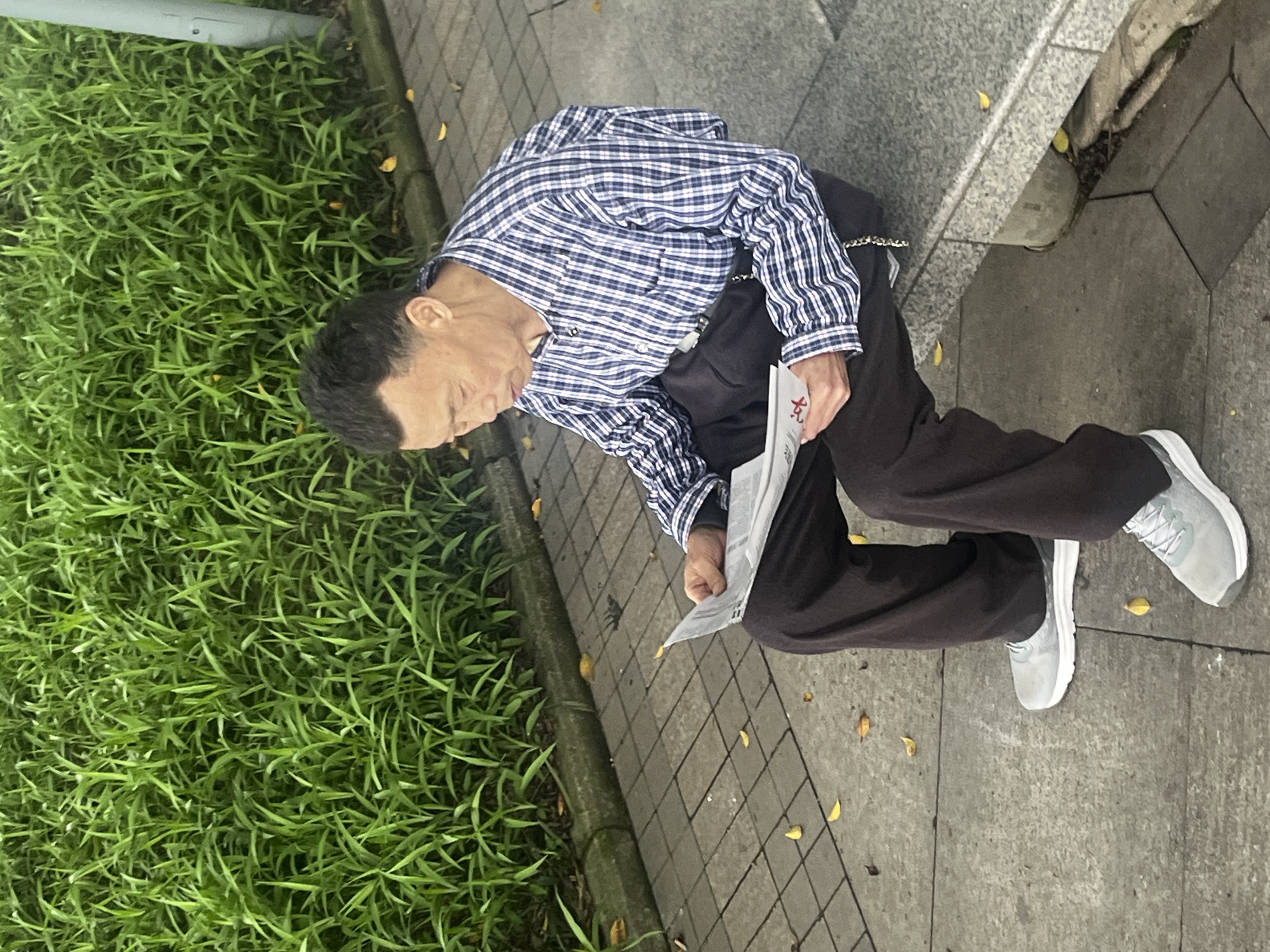 An elderly Chinese man is seated on the edge of a concrete structure next to a low concrete wall bordering a patch of lush green vegetation, reading a newspaper. He is wearing a blue and white checkered long-sleeved shirt, dark pants, and gray athletic shoes. The man is positioned on a paved walkway made of square concrete tiles. Scattered yellow leaves are visible on the walkway. The location is Jiangjin, China.
