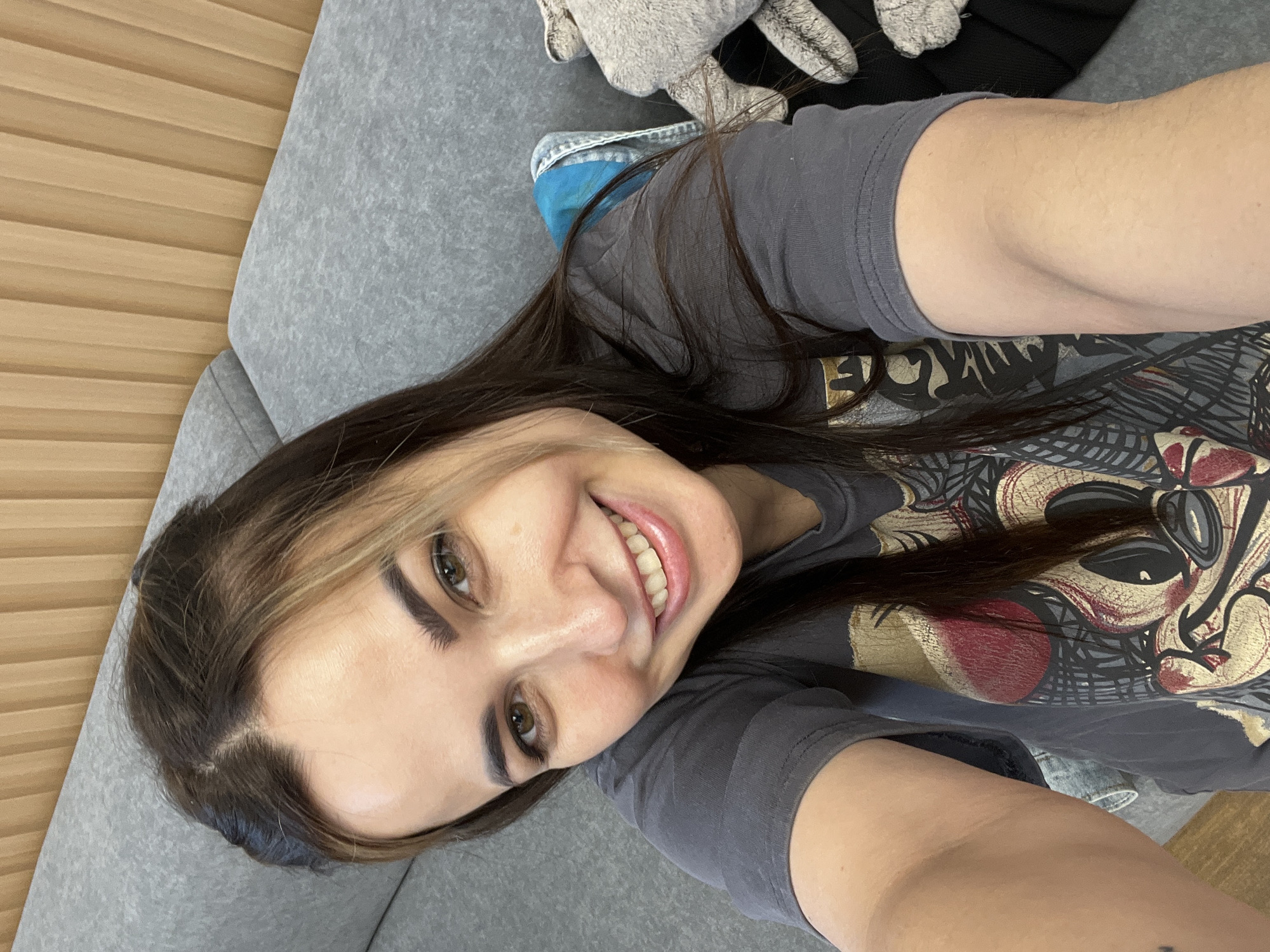 A young woman with long, dark hair and light brown highlights smiles at the camera, holding it up in a selfie. She is wearing a dark gray t-shirt with a cartoonish graphic on the front. She is sitting on a gray sofa with a light wooden wall behind it. There is a bundle of gray, fuzzy fabric to her right.