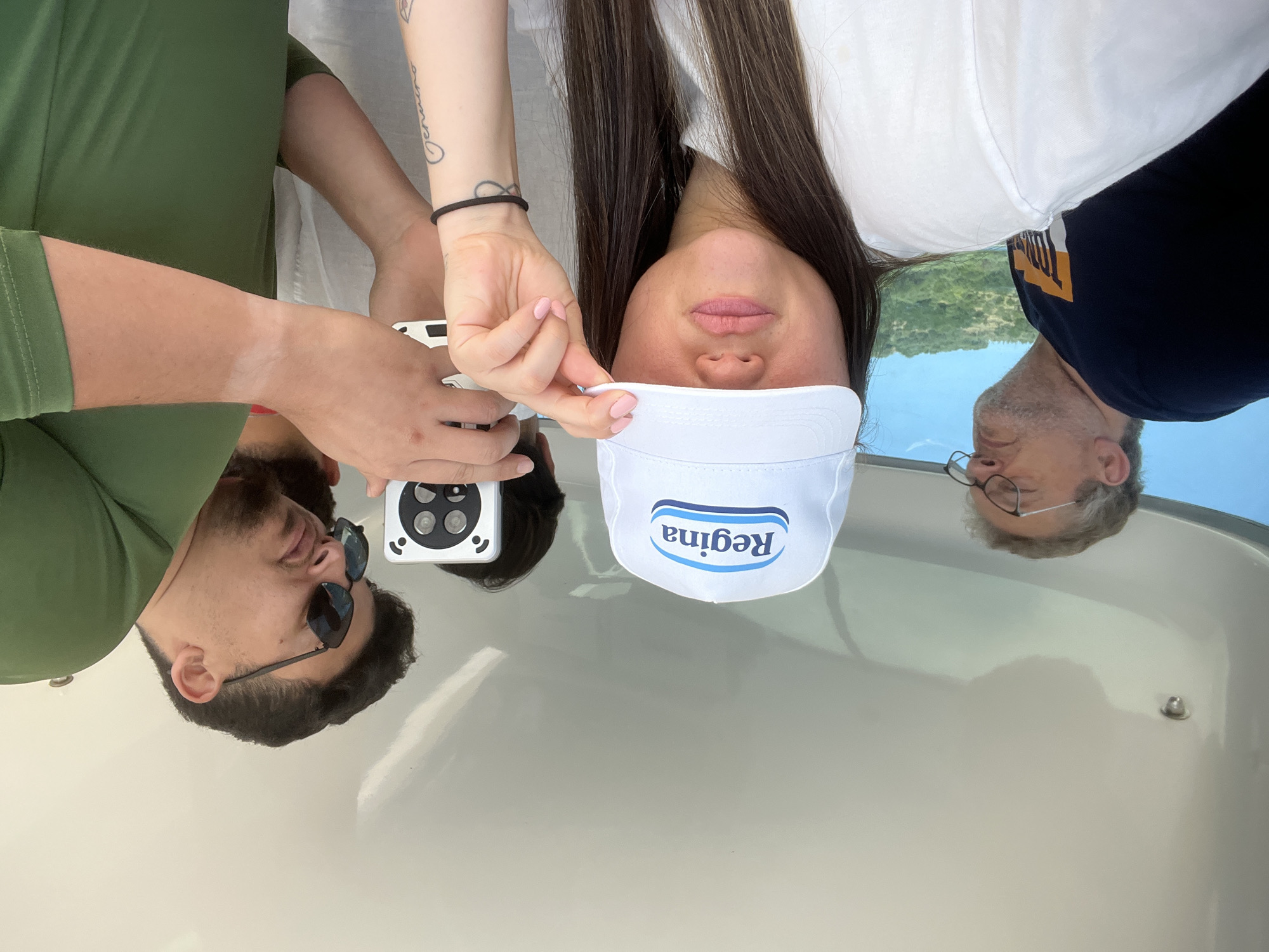 This image captures a creative group portrait featuring three individuals, presented as reflections on a glossy, light-colored surface, likely the deck of a boat. The reflections show the people appearing upside down against a backdrop of a bright blue sky and lush green hills, hinting at a sunny day in a scenic coastal or lakeside setting, consistent with Pollica, Italy.
In the center of the reflection is a woman with long dark hair, wearing a white top. She is holding a white visor with both hands, her light pink fingernails visible. The visor clearly displays the word "Regina" in blue letters, though it appears reversed due to the reflection. A tattoo with script is visible on her right forearm.
To the left, a man with dark hair and a beard, wearing a green top and sunglasses, is also reflected. He is holding a white device with black circular features, possibly a phone or camera, suggesting he might be capturing the scene or participating in the photo.
To the right, an older man with grey hair and glasses, dressed in a dark blue shirt with a yellow and orange graphic, completes the trio.
The image utilizes the reflective surface to create an inverted and artistic perspective of the subjects. The bright illumination indicates it is daytime, and the relaxed postures suggest a casual outdoor activity.