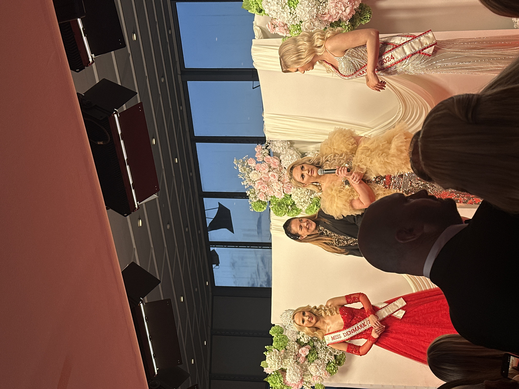 A stage is set with several women standing on it. One woman is wearing a red gown and a sash that reads "MISS DENMARK". Another woman is wearing a sparkling dress and a sash labeled "2024". A third woman holds a microphone and is wearing a beige dress with a feathered boa. A fourth woman is also present on the stage. The stage backdrop features white fabric and floral arrangements. The background reveals a modern building with large windows, suggesting an indoor event. The location is Skovshoved, Denmark, as implied by the sash identifying "MISS DENMARK". Attendees are visible in the foreground, including a man in a suit.
