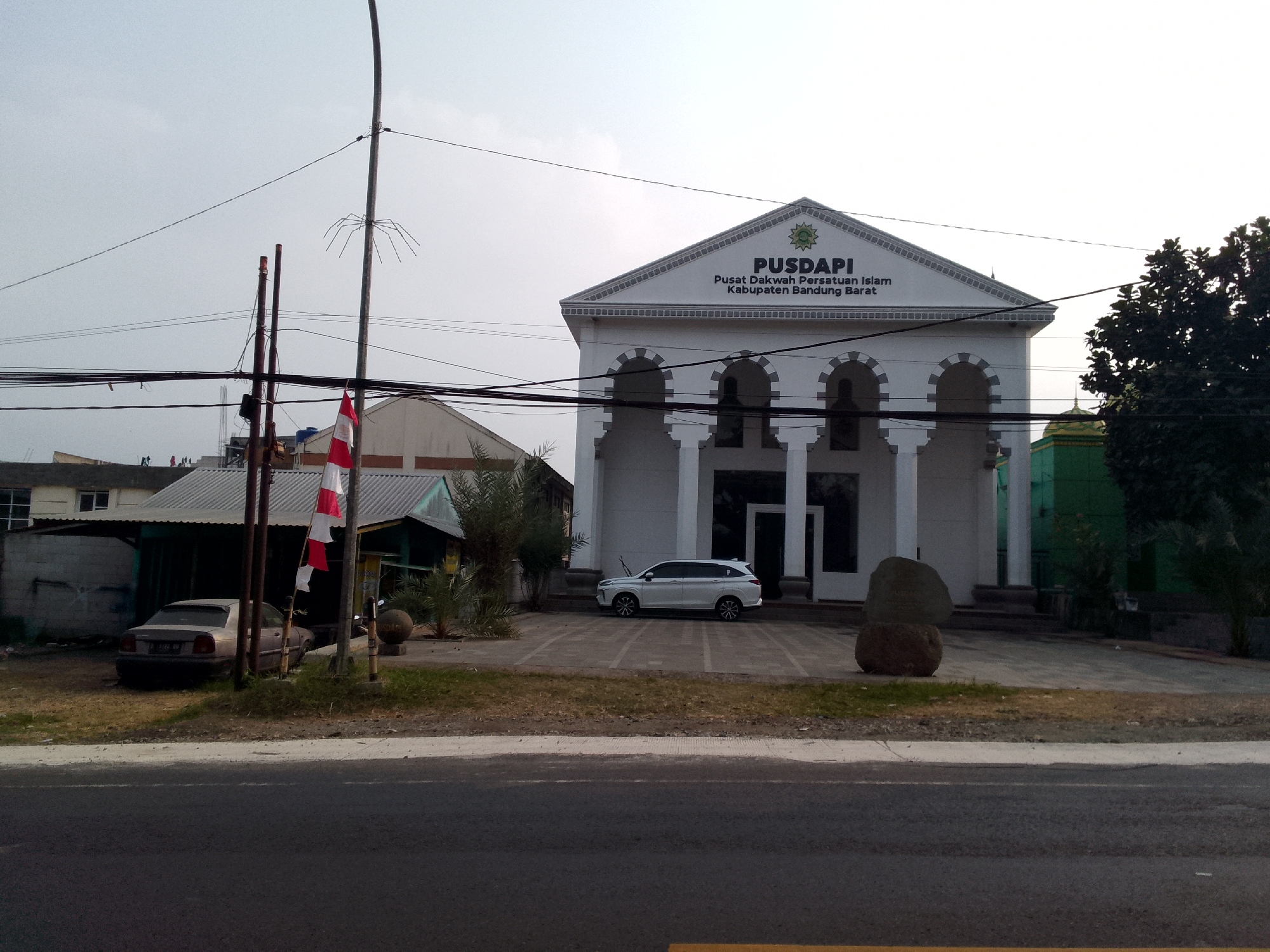This is a full outdoor shot of the PUSAPI building, identified as "Pusat Dakwah Persatuan Islam Kabupaten Bandung Barat" in Mandalasari, Indonesia. The building is white with a prominent triangular pediment that displays the name of the organization in dark lettering. Large, arched windows and white columns line the facade. In front of the building, a white SUV is parked in a designated space. To the left of the building, a smaller, low-roofed structure is visible, with a silver car parked in front of it. Red and white Indonesian flags are displayed on a pole in front of the smaller structure. The scene is bordered by a road in the foreground and a clear sky above, with some power lines crisscrossing the frame. Lush green trees and foliage are also present on the right side of the image, with a green domed structure peeking through them. The ground is covered in dry grass and scattered rocks. The overall lighting suggests it is daytime. There are no people visible in the frame.
