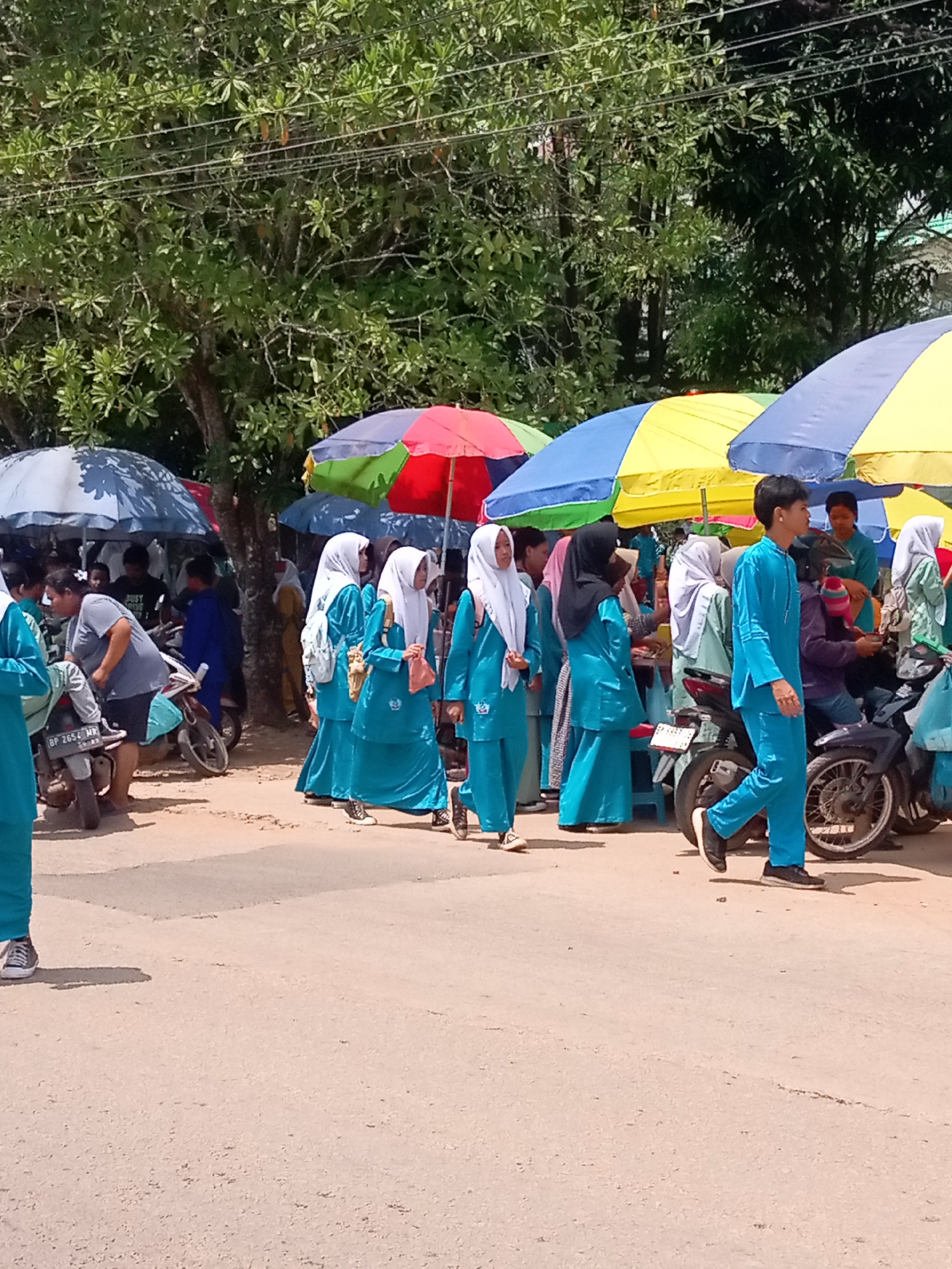 A group of people are gathered outdoors, with several individuals wearing teal Islamic school uniforms. Some of the uniformed individuals are walking and conversing, while others appear to be waiting or observing. There are several motorcycles parked in the vicinity. Large, colorful umbrellas, in shades of blue, yellow, red, and green, provide shade over stalls or gathering areas. Trees with green foliage are visible in the background, suggesting an outdoor market or event. The ground is a mix of paved road and dirt. The scene appears to be taking place in Batam, Indonesia, likely in an area where such community gatherings or events are common.