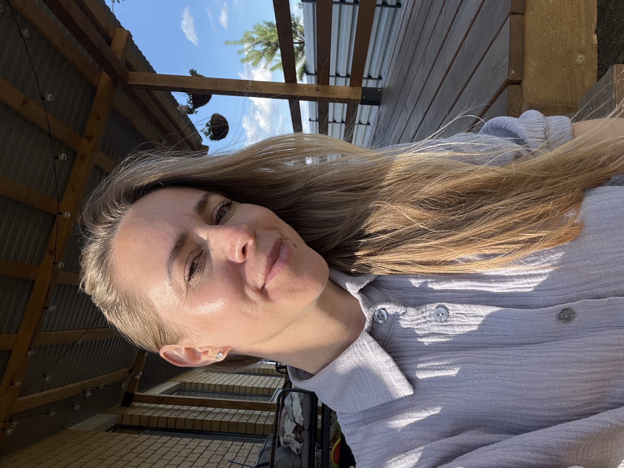 This media file shows a close-up, sideways selfie of a woman with long, blonde hair. She is wearing a light purple, crinkled button-up shirt. She has light skin, light brown eyes, and is smiling subtly. She has a small silver earring in her left ear. The background is an outdoor setting with wooden planks and a corrugated metal roof structure. There are hanging plants visible, and the sky is blue with some white clouds, suggesting daytime. The lighting indicates it might be late afternoon or early morning, with sunlight casting shadows. The woman appears relaxed and happy. The setting appears to be a porch or an outdoor covered area. No text is visible.