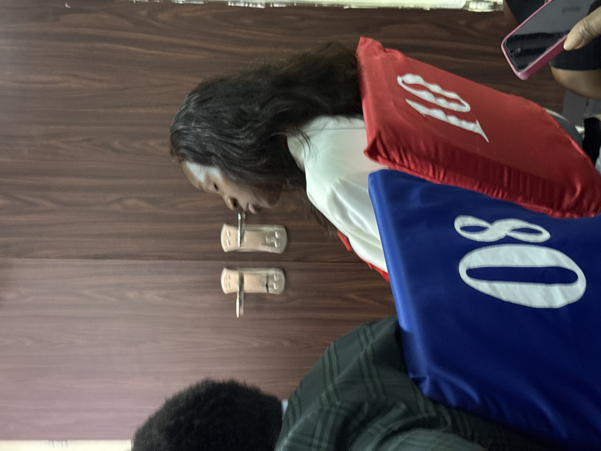 In a room with dark wood-paneled doors and two silver door handles, a woman with long, dark hair is seated in a red chair. The chair has a white number "18" embroidered on it. To her left, another person is partially visible, wearing a green and black plaid jacket and seated in a blue chair with a white number "08" embroidered on it. The woman in the red chair appears to be sleeping or resting with her eyes closed. In the lower right corner, a hand is holding a pink-cased smartphone. The scene is indoors, likely in a formal or semi-formal setting.