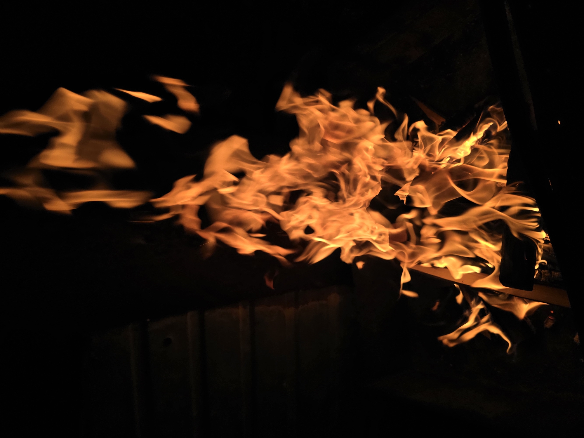 This is a close-up photograph of a campfire in the dark, with flames dominating the center of the frame. The flames are a vibrant orange and yellow, with some areas appearing white-hot. The background is predominantly black, suggesting the photograph was taken at night or in a dimly lit environment. To the left of the flames, there is a dark, textured surface that appears to be made of wood, possibly the side of a fire pit or structure. There are also dark, indistinct shapes at the bottom of the frame, which are likely the burning logs. The overall impression is one of intense heat and light emanating from the fire, contrasted with the surrounding darkness. There are no people or animals visible in the image, and no context is provided regarding the location in Checheleve, Ukraine.