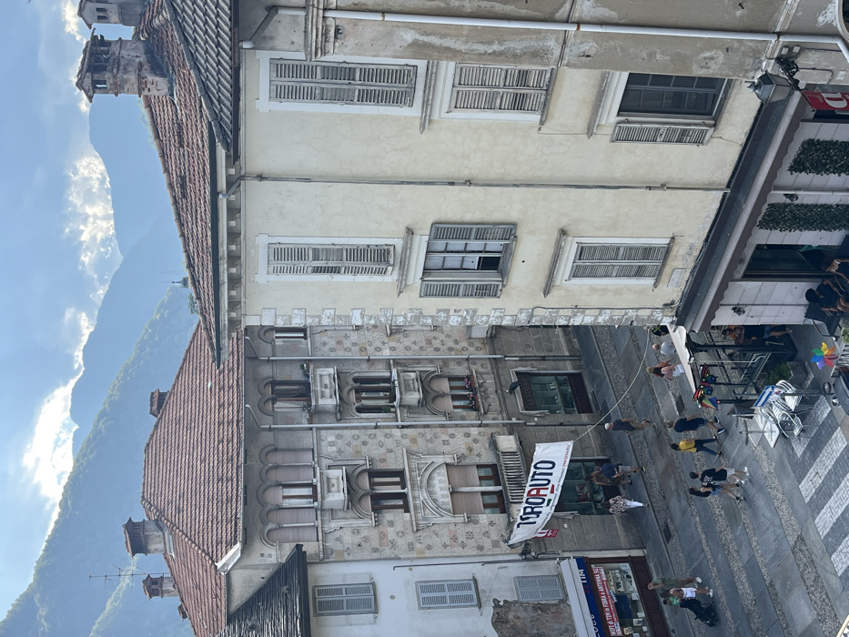 This elevated view captures a daytime street scene in Varallo, Italy. In the foreground on the right, a multi-story building with a pale yellow stucco facade features multiple windows, predominantly fitted with closed, weathered light-colored wooden shutters; one window on the mid-level is open. The ground floor of this structure contains a commercial establishment, identifiable by its outdoor seating area with stacked chairs and tables, an overhead awning, and decorative green wall panels.
Across the street, a more ornate multi-story building displays a patterned light-colored stone facade with decorative elements, including arches and small balconies. A large white banner with "TOROAUTO" in blue and red text is suspended from a balcony railing on this building. To the far left, a portion of a simpler white building is visible, featuring dark shutters and a shopfront displaying "TIM" and "5G" signage.
The street below is paved with grey pavers, exhibiting a subtle diagonal pattern, and includes a white-striped crosswalk in the immediate foreground. Several pedestrians populate the street: on the left, an individual pushes a baby stroller; in the center, a group of approximately eight individuals walk from left to right; and near the right-hand commercial establishment, several people are seated at tables or standing. A light-colored rope or cable stretches horizontally across the street at pedestrian height, originating from the building on the right.
The background features hazy, blue-green mountains beneath a partly cloudy sky. All visible buildings are topped with red terracotta tile roofs and feature brick chimneys.