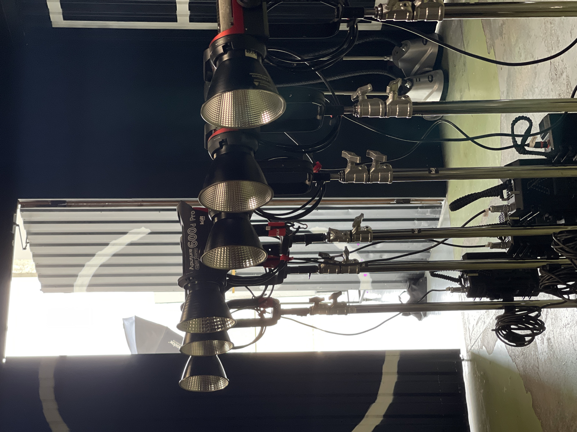 The image shows a close-up, eye-level view of several professional film lighting fixtures, specifically Aputure 600d Pro lights, positioned in a row. These lights are mounted on tall, silver stands and are angled towards the left side of the frame, where a large open doorway floods the scene with bright, natural light. The background consists of a dark, studio-like environment with corrugated metal walls and some industrial equipment, including what appear to be power supplies or ballasts connected to the lights. The floor is a light-colored, possibly concrete, surface with some puddles of water. The overall impression is that of a film or photography studio set. There are no people visible in the image, and no specific details suggest a location in Kyiv, Ukraine.