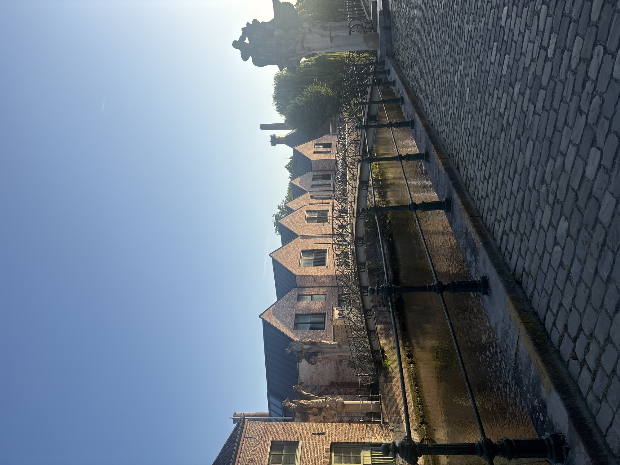 The photograph captures a serene canal scene in Ghent, Belgium, likely taken during the early morning given the soft light and clear sky. The left side of the image features a row of historic brick buildings with dark-tiled roofs and visible chimneys. Ornate statues stand in front of the buildings, adding a touch of classical artistry to the urban landscape. A decorative wrought-iron bridge spans the tranquil canal, its reflection subtly visible in the water. The right side of the image is dominated by a cobblestone path leading to a large, abstract stone sculpture. The canal is bordered by a dark green metal railing, and the water is calm, reflecting the sky and surrounding architecture. Trees and greenery are visible in the background, particularly on the right side, hinting at a park or natural area. The overall impression is one of historical charm and peaceful urban beauty.