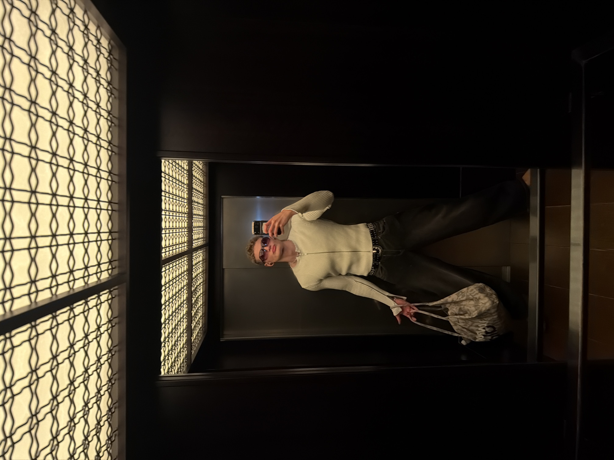 A young man is taking a selfie in an elevator. The man is wearing a white zip-up sweater, black pants, and red-tinted sunglasses. He is holding a patterned tote bag in his left hand and a smartphone in his right. The elevator has decorative patterned walls, and the overall ambiance is dark with patterned light behind. The location is an elevator in the city of London, United Kingdom.