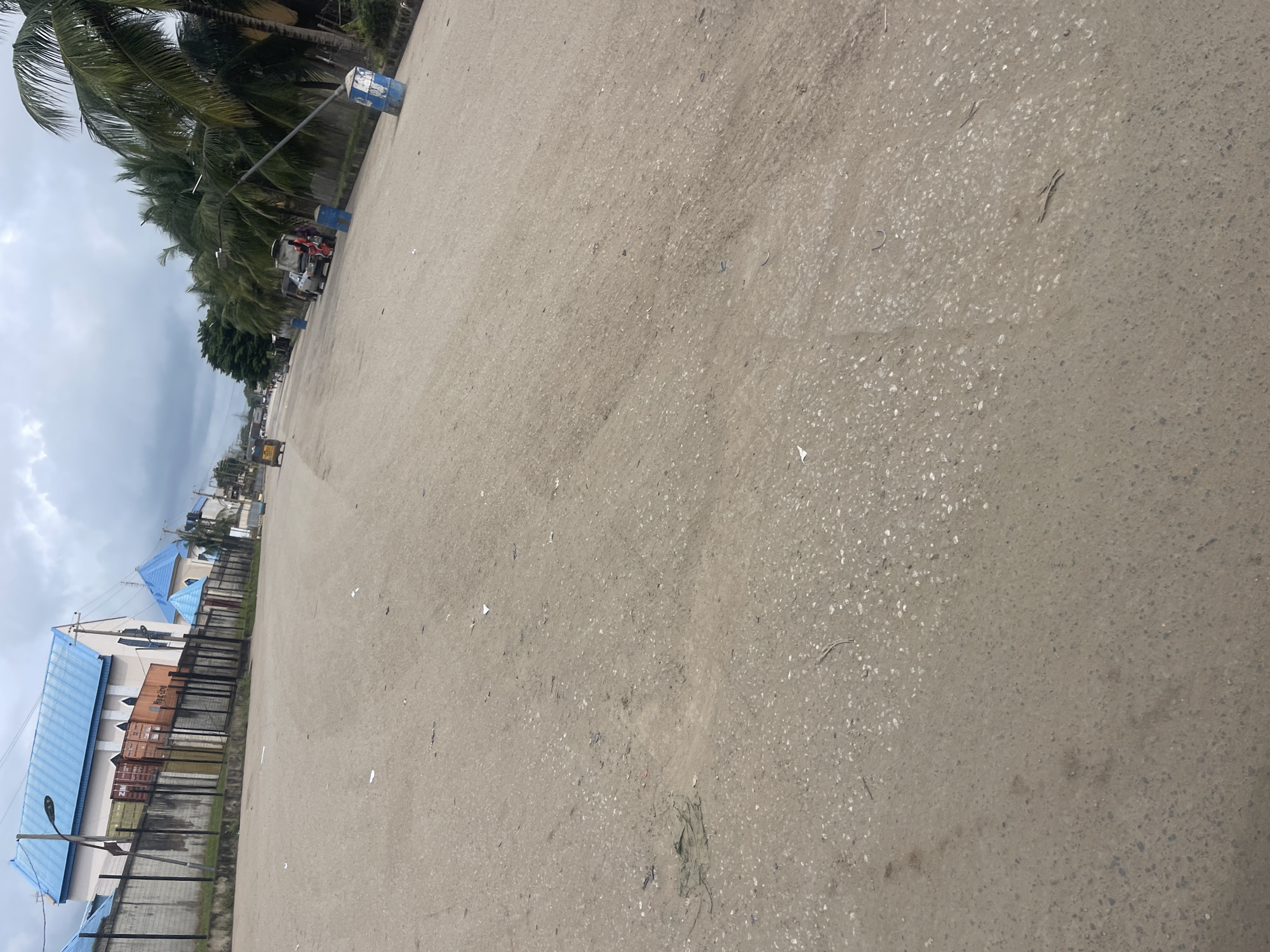 This is a medium-angle, eye-level outdoor shot of a paved road in Onitsha, Nigeria. The road appears to be a main thoroughfare, with two lanes visible. The surface of the road is cracked and uneven, with patches of dirt and debris scattered across it. On the left side of the road, there is a wall with a fence, behind which are buildings with blue roofs. Several shipping containers are stacked behind the wall. On the right side of the road, palm trees line the street. Several vehicles are present, including a car, a trishaw, and possibly a truck in the distance. Two blue trash bins are also visible along the roadside. The sky is overcast, suggesting cloudy weather. The overall impression is of a busy, somewhat aged urban environment.