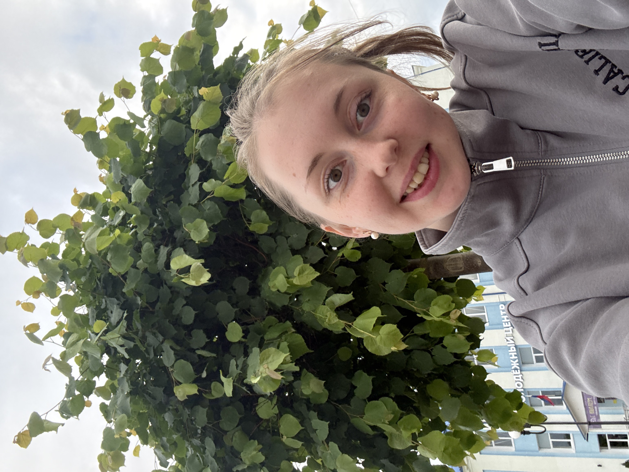 An outdoor selfie captures a young adult woman smiling in the foreground, with a large deciduous tree and a building in the background. The woman has fair skin, light brown hair partially tied back, and light brown eyes. She displays a broad smile, revealing her teeth, and wears small, light-colored stud earrings. Her attire consists of a grey zippered jacket, with a silver-toned zipper pull and visible embroidery reading "CALIF" on her right side. Behind her, a full, green-leaved tree dominates the midground; some leaves exhibit a yellowish tint, and a portion of its brown trunk is visible on the right. In the far background, a multi-story building painted light blue and white features multiple windows. White Cyrillic text, partially obscured, reads "ОДЕЖНЫЙ ЦЕНТР" (Youth Center) on a blue band, with "С ЮБИЛЕЕМ" (Happy Anniversary/Jubilee) below. A flagpole with a red, white, and blue striped flag is positioned in front of the building, and a black streetlight stands to the left. The sky is overcast and light grey. The setting indicates an urban or semi-urban environment in the city of Unknown, Unknown.