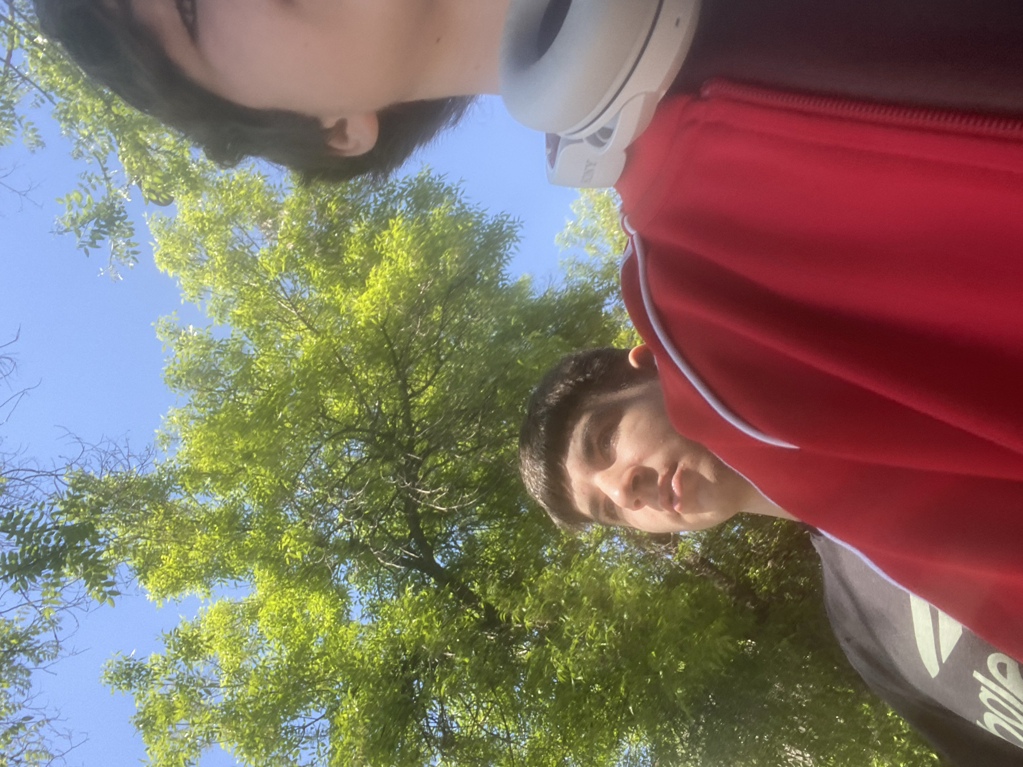 Two individuals are captured in a selfie-style photograph taken outdoors. The perspective is from below, looking up toward the subjects and the sky. The top individual is partially visible, showing a fragment of a face, dark hair, and white headphones around the neck. The bottom individual is more clearly visible, with a complete face and dark hair. A red jacket is draped across the right side of the frame. Green leafy trees occupy the middle ground of the frame, with blue sky visible between the branches. The location is not discernible from the given image. The city of Plovdiv, Bulgaria cannot be definitively identified as the setting.