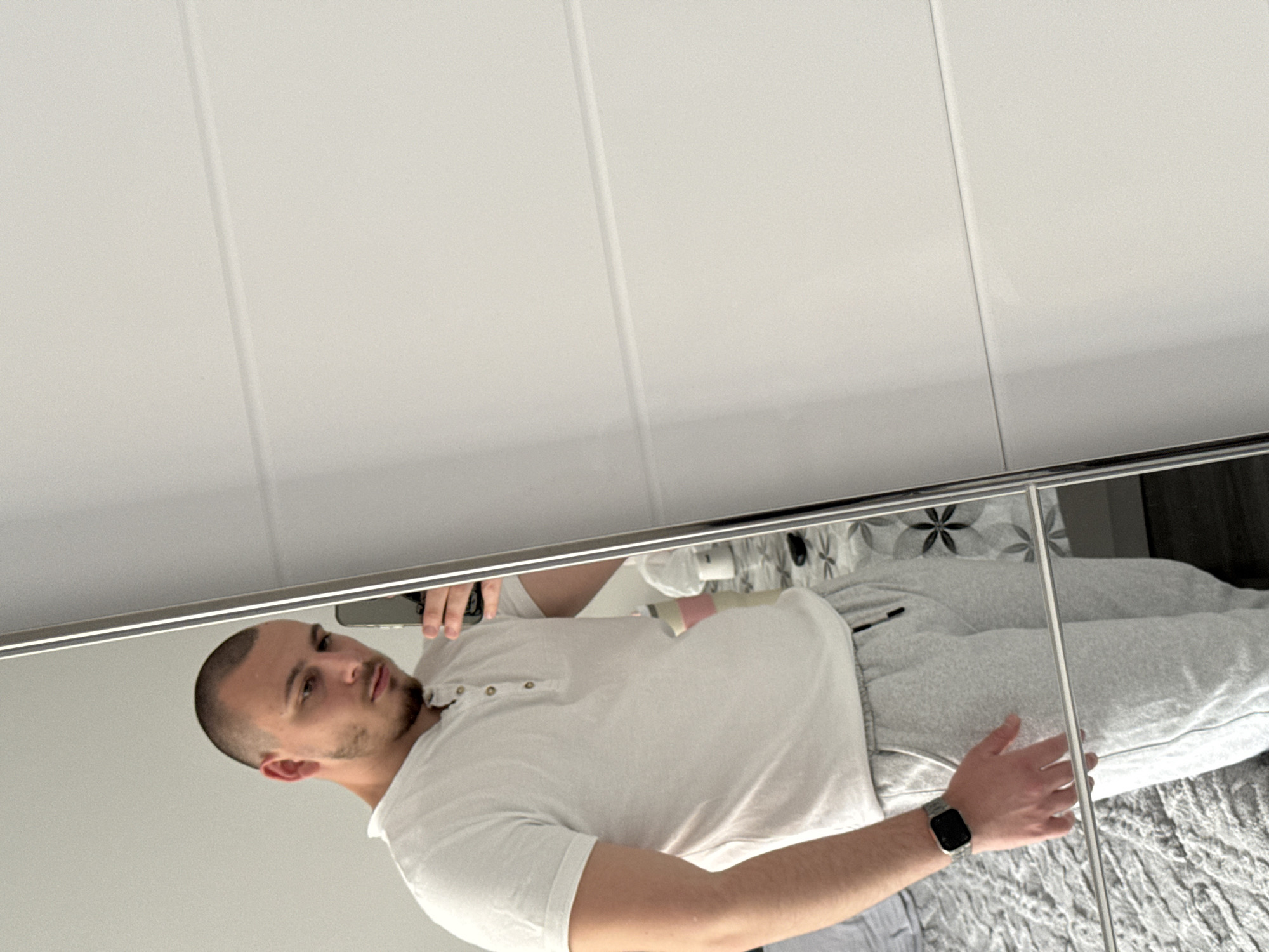 A man is taking a selfie in a mirrored sliding door wardrobe. He is wearing a white short-sleeved shirt and gray sweatpants. He has short, dark hair and a beard. He is wearing a smart watch on his left wrist. The wardrobe has three mirrored doors and is located indoors, likely in a bedroom. The location in the city of Stara Zagora, Bulgaria is unknown.