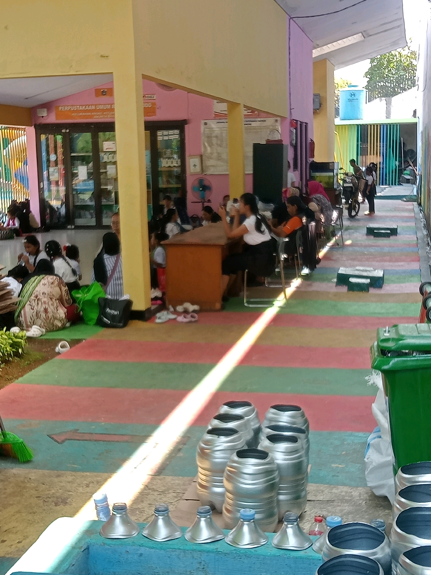 This image captures a scene at the Perpustakaan Umum R.A. Kartini, a public library in North Jakarta, Indonesia. In the foreground, a collection of various sized cylindrical silver containers with black openings are arranged on a blue surface, with some smaller silver, funnel-shaped objects and a few plastic bottles near them. To the left, a green trash bin is visible. The ground is covered with a colorful striped mat in shades of red, green, and blue. A broom with green bristles rests on the ground near the mat.
In the middle ground, a group of people, primarily women and children, are seated or standing. Several individuals are gathered around a wooden desk, some appearing to be engaged in activities. Others are seated on chairs or on the colorful mat. Behind them, the exterior of the library is visible, featuring large glass windows and doors. A sign above the entrance reads "PERPUSTAKAAN UMUM R" and partially visible text below it. The building is painted in pastel colors, including yellow and pink.
In the background, more people are visible, including some standing near a motorcycle. A blue water tank is mounted on a structure. The scene appears to be outdoors, possibly a courtyard or an open area adjacent to the library. The overall impression is of a community gathering or an event taking place at the library.