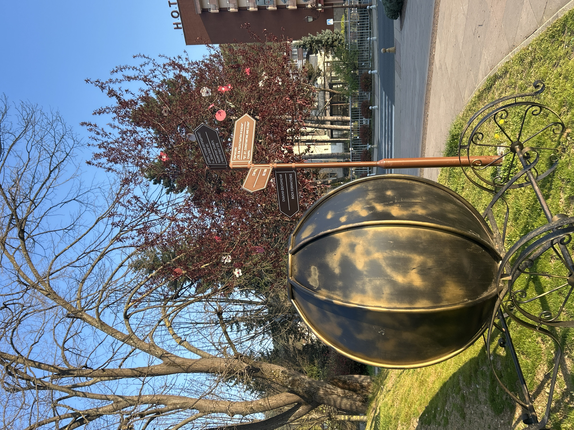The scene depicts a black and gold metallic sculpture resembling a bicycle cart with a large spherical container, located in Zlatograd, Bulgaria. A directional signpost with multiple arrow-shaped signs is attached to the structure. The signs provide directions to "Old Town", "Assumption Church, 1 Penyo Penev str", "ST. ST. Constantine and Elena Chapel", and "Rock Formations" in both Bulgarian and English. The sculpture sits on a grassy area adjacent to a pedestrian crosswalk and a street with traffic calming bollards. In the background, there is a deciduous tree with red leaves and a building labeled "HOT". A bare tree is also partially visible in the lower left corner. The sky is blue and clear.