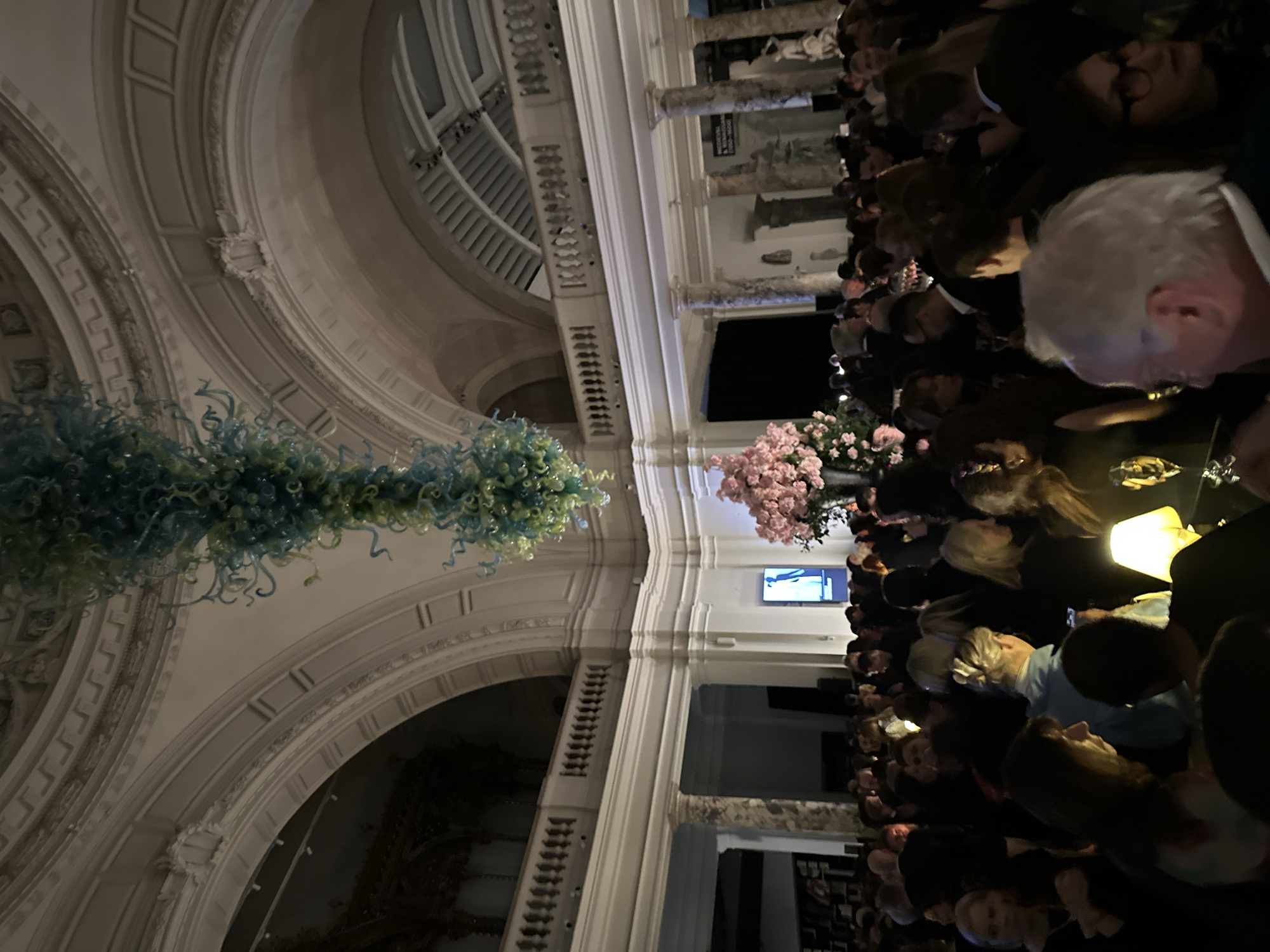 The image depicts a crowded evening event occurring within the Victoria and Albert Museum (V&A) in South Kensington, London, United Kingdom. The setting is a large, ornate hall featuring high vaulted ceilings, multiple architectural arches with decorative molding, and an upper-level balcony with a white balustrade. Dominating the central overhead space is a large, intricate glass sculpture composed of numerous blue and green organic forms, characteristic of Dale Chihuly's work.
The lower floor of the hall is densely populated by approximately 100-150 individuals, primarily adults, most standing and engaged in conversations. Attendees are dressed predominantly in dark, semi-formal attire. Several individuals are visible holding drinks, including champagne flutes. A prominent feature on the right side of the image is a large floral arrangement consisting of numerous pink flowers, likely peonies or hydrangeas, displayed in a dark, vase-like container. In the background, white marble columns, classical statues, and relief panels are discernible, along with a tall, illuminated digital display screen exhibiting a light-colored image. Lighting is dim and ambient, with localized illumination from unseen sources and a small, glowing table lamp in the bottom-center foreground. The scene presents a lively social gathering or reception within a grand institutional interior.