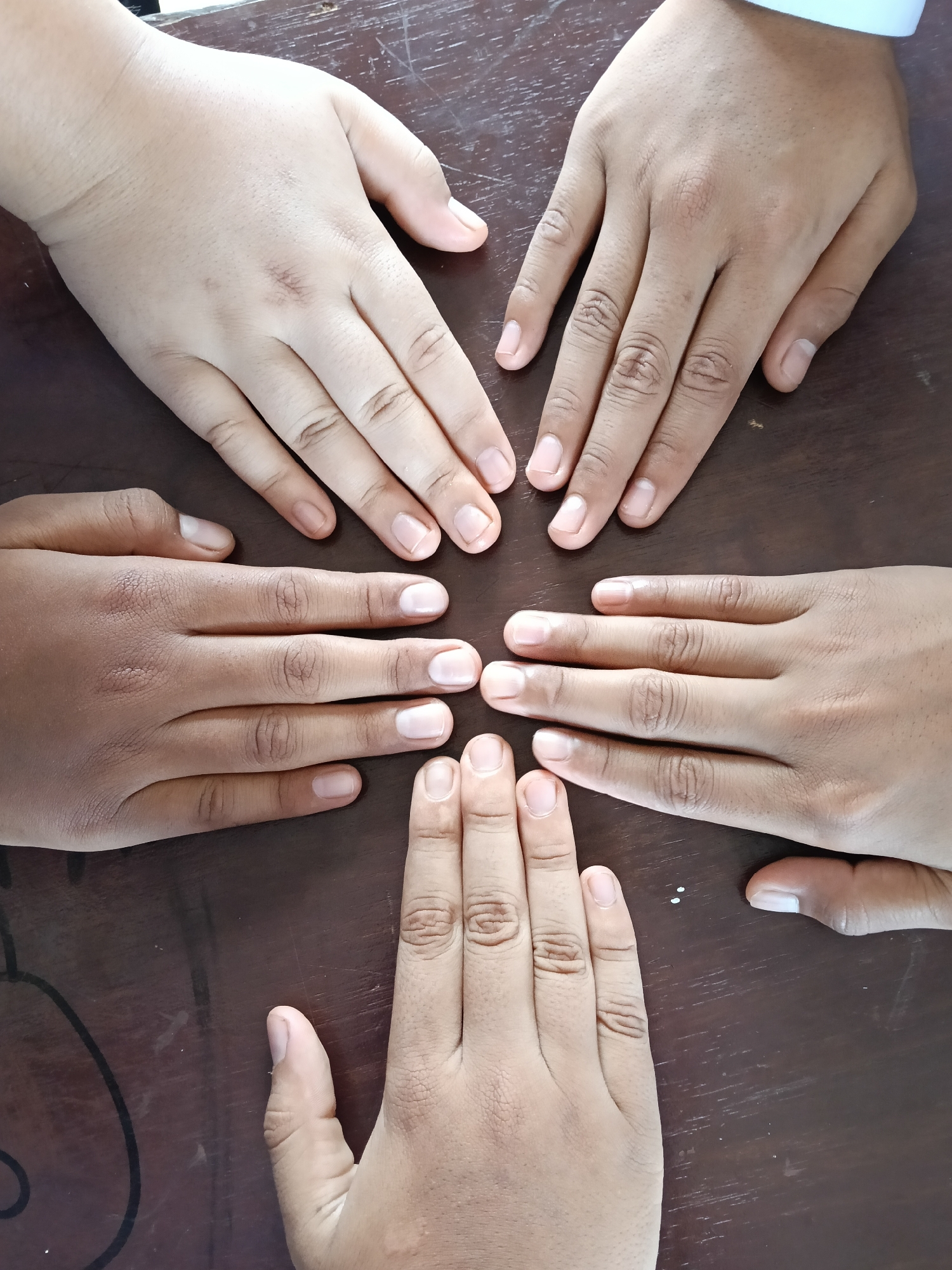 Four human hands of varying skin tones are arranged on a dark brown, possibly wooden, surface. The hands are positioned to point towards a central point, with the tips of the fingers almost touching. The fingernails are short and unpolished. The background appears to be a simple, unadorned surface. There are no visible markers to identify the location as Lhokseumawe, Indonesia.