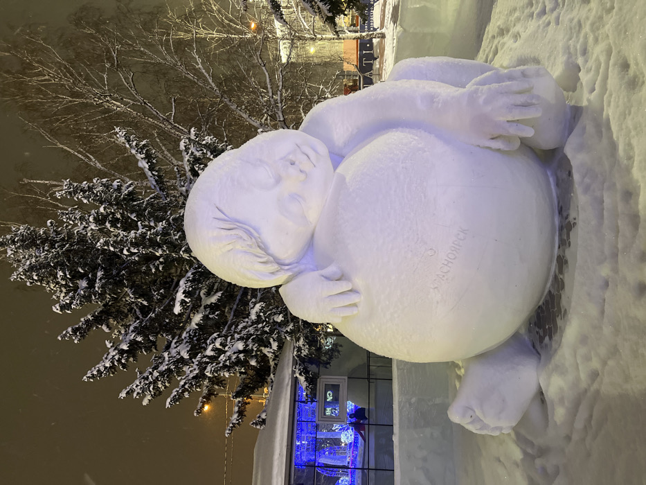 A large, white snow sculpture depicting a sleeping, cherubic figure is prominently featured in an outdoor urban setting during nighttime in Krasnoyarsk, Russia. The figure is seated, leaning its head with closed eyes on a large, rounded belly or spherical form. Its left arm is draped over this central mass, while its right arm rests alongside its body. The word "КРАСНОЯРСК" is carved into the front surface of the sculpture's belly.
The ground around the sculpture is covered in fresh snow. In the background to the left, a modern building with large glass windows is visible, illuminated by blue decorative string lights. Reflections in the windows suggest interior elements and a distant human figure. An ice block structure stands between the sculpture and the building. Further into the background, a dense evergreen tree, heavily laden with snow, stands beside bare deciduous trees, also covered in snow. Distant streetlights emit a warm orange glow, and illuminated signs with Cyrillic text (e.g., "ATI") are visible on other structures. The sky is dark, indicating night, and light precipitation, consistent with snow or sleet, is visible in the air. The sculpture itself is illuminated, exhibiting subtle purple and blue hues.