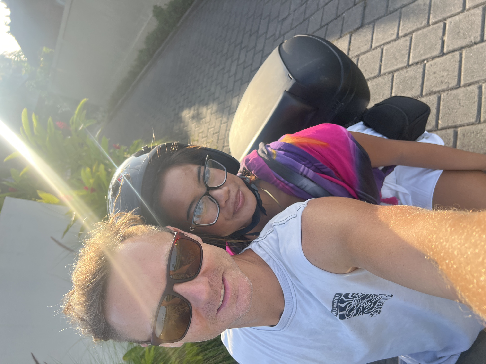 A couple is posing for a selfie on a scooter in Ubud District, Indonesia. The man is wearing sunglasses and a white t-shirt, while the woman has a helmet on and is wearing glasses. They are in front of a dark colored scooter with a top case and a small seat, riding on a stone-tiled street. The background features lush greenery and a building partially visible with sunlight.