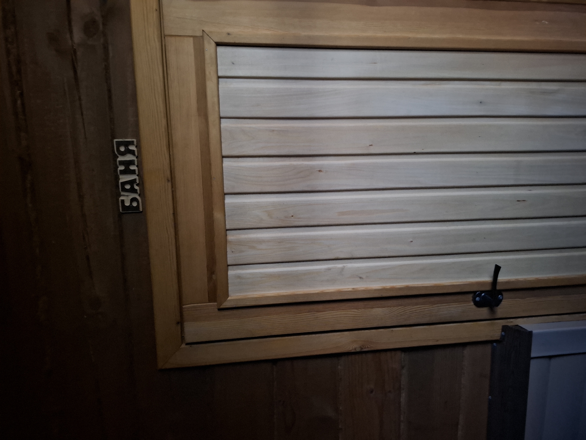 The image shows a wooden structure, likely a window or vent, inside a room. The structure is made of horizontal planks, light in color, framed by a darker, more textured wood. To the left of the structure, the Russian word "БАНЯ" (Banya, meaning bathhouse) is spelled out vertically in individual letters. The room's walls are constructed of wooden planks, also varying in tone and texture. A black metal latch is visible on the lower right of the wooden structure. A portion of another structure, possibly a bench or paneling, is visible at the lower right corner. The setting appears to be the interior of a sauna or bathhouse in Амзя, Russia.