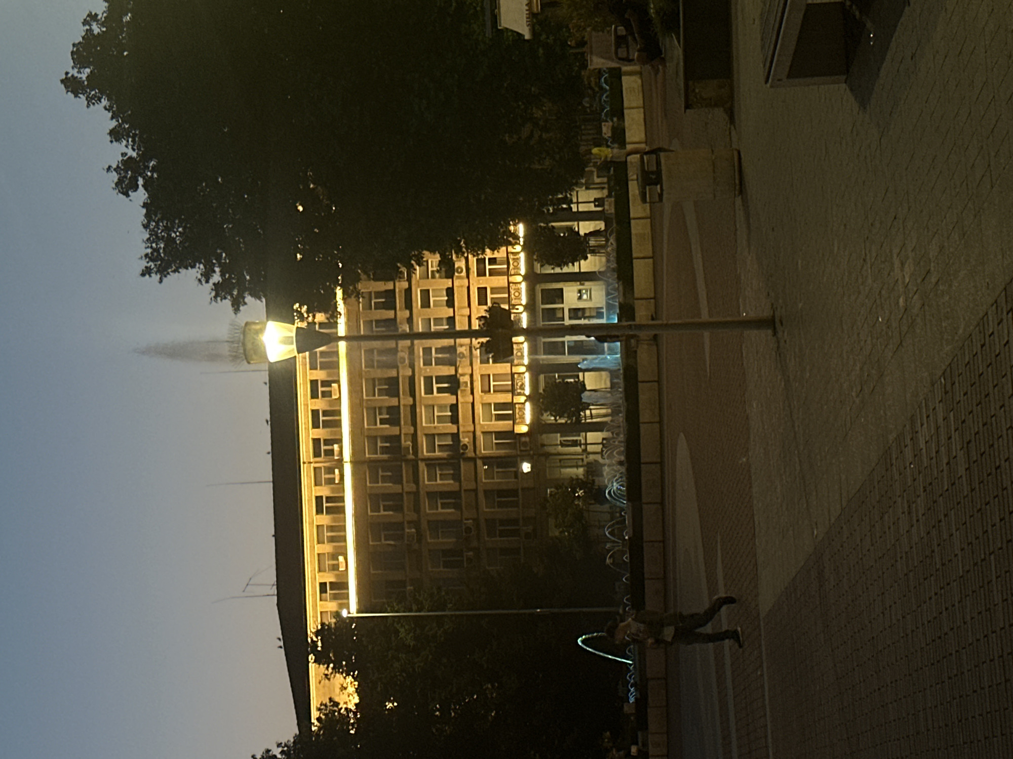 In an outdoor urban setting at dusk, a building with multiple stories and numerous illuminated windows is visible. The building is lit with warm, yellow light, highlighting its architectural features. In front of the building, a fountain displays jets of water illuminated with blue and green lights. A single street lamp stands in the foreground, its light illuminating the paved ground. A person, dressed in casual clothing, walks across the paved area, and a trash receptacle is situated to the right of the street lamp. Trees are present on both sides of the scene, with foliage obscuring parts of the building and the sky. The sky is a gradient of dark blue to light blue, indicating twilight.