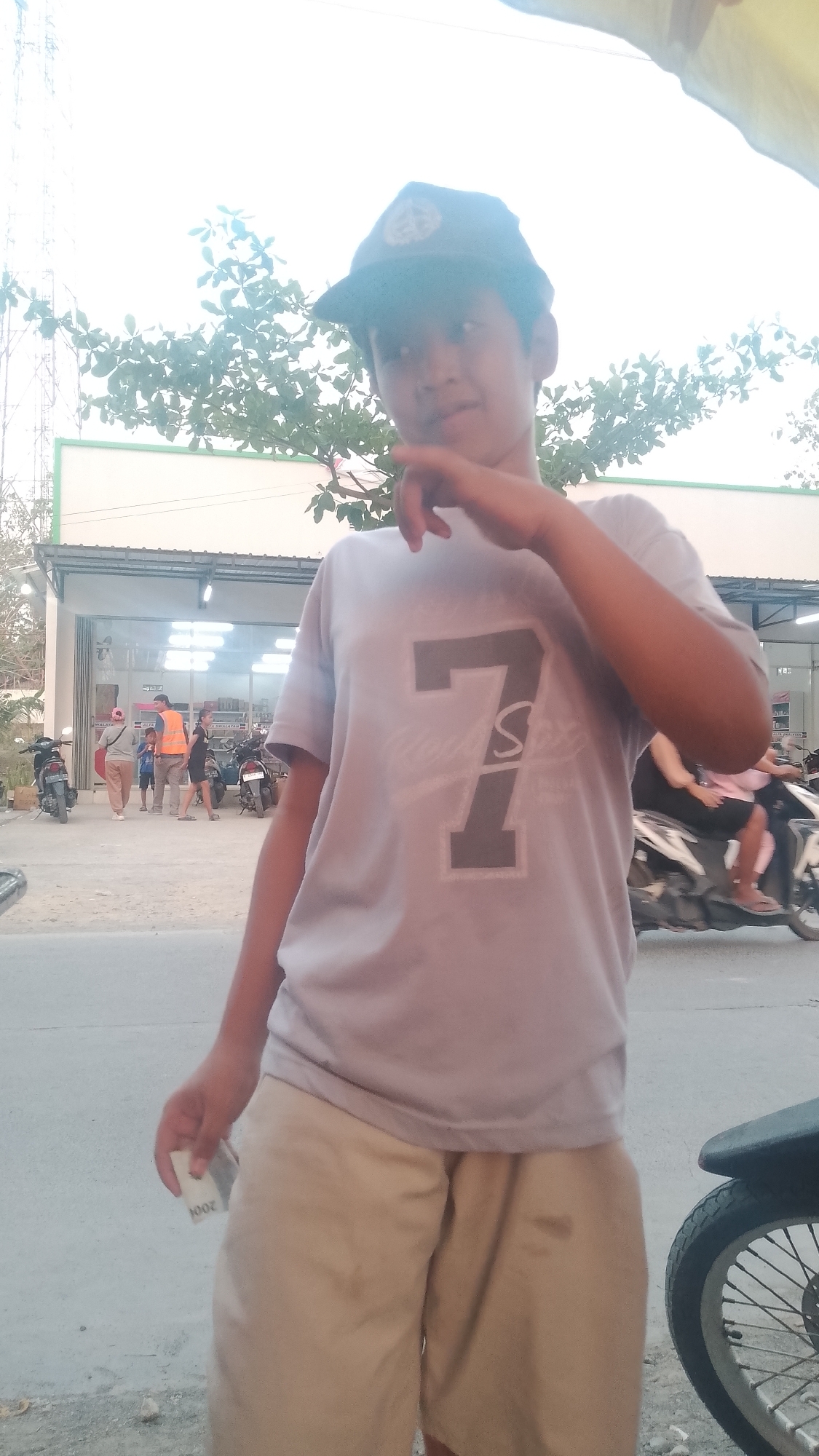 The image depicts a young male wearing a light purple t-shirt with a large grey number "7" and some text below it, and light beige shorts. He is also wearing a dark baseball cap. The individual is standing outdoors, with his right hand near his mouth and his left hand holding a small white object, possibly a piece of paper or a bill. In the background, there is a building with a green trim and large glass windows, which appears to be a store or commercial establishment. Several motorcycles are parked in front of the building, and a few other individuals are visible in the distance, some walking and some standing. Trees with green leaves are present, and a tall tower structure is partially visible in the upper left portion of the frame. The ground is paved, suggesting an outdoor area in a developed setting. The overall scene suggests a casual outdoor environment, possibly a street or parking area in a town or city. This location is identified as Karanglangu, Indonesia.
