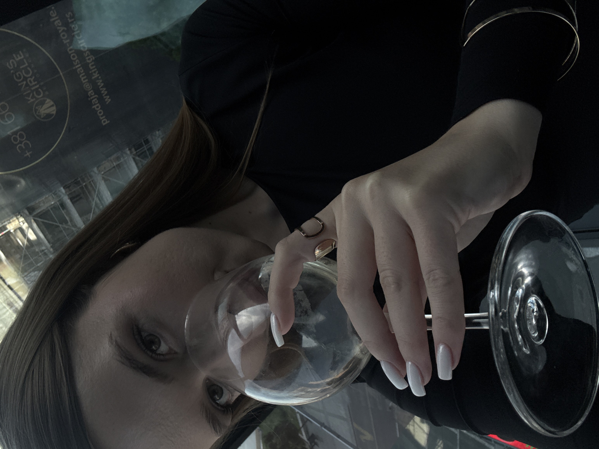 A rotated, close-up image shows a woman holding a wine glass. She has light skin, brown hair and wears gold rings and bracelets. The glass is partially filled with a clear liquid. A reflection is visible on the glass, featuring the contact information "prodaja@maison-royale" and the website "www.kingscircle.rs". This indicates the location might be related to King's Circle in Vracar Urban Municipality, Serbia.
