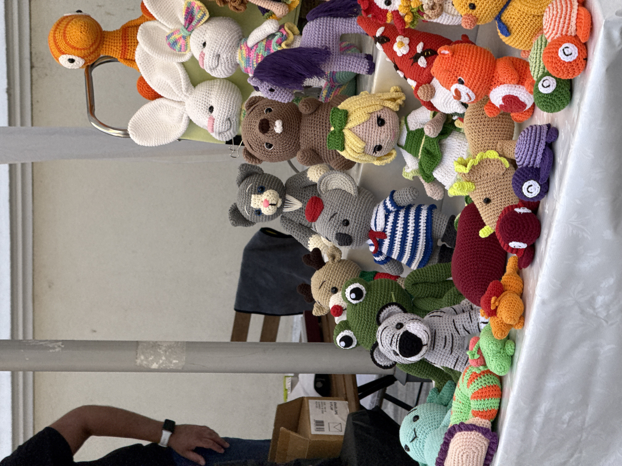This image depicts a close-up, eye-level view of a table laden with a variety of crocheted stuffed toys, likely displayed at an outdoor market. The toys are diverse in form and color, including animals like a frog, a tiger, a bear, a koala, a deer, a horse, and bunnies, as well as a doll and an orange, alien-like figure. The crocheted items exhibit intricate patterns and textures. In the background, a person's arm and torso are visible on the left, wearing a black t-shirt and a watch. Behind the toys, a wooden chair with a dark fabric draped over it is partially visible, along with a white vertical pole and a beige wall. The table is covered with a white, textured cloth. The lighting suggests an overcast or shaded environment. The specific location of Bansko, Bulgaria, is not directly discernible from the visual elements within the image.