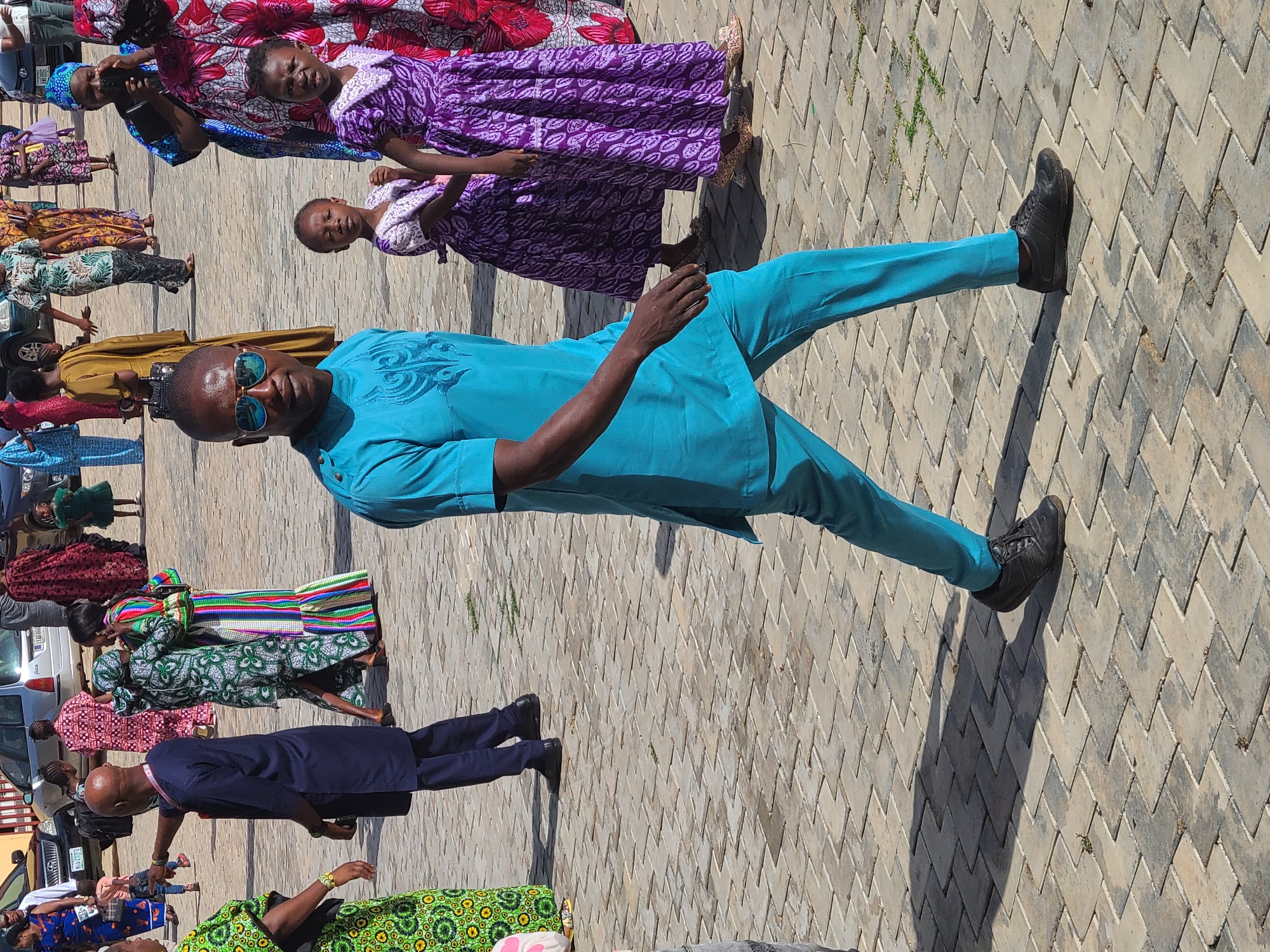 The image shows a man wearing a bright blue outfit and sunglasses, standing on a paved surface. There are other people in the background, some dressed in traditional African clothing. The location appears to be an outdoor area in a city. The city and precise location are not specified.