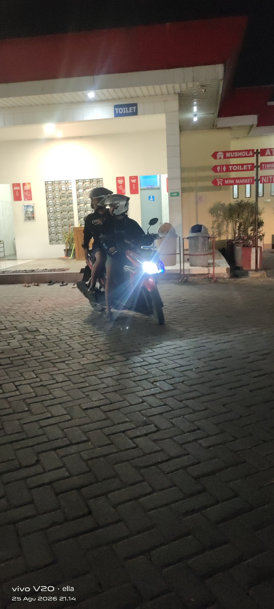 The image captures an outdoor scene at night, likely in a public area with paved brick flooring. Two individuals wearing helmets are seated on a red motorcycle, which has its headlight on. The motorcycle is positioned on the paved ground, facing forward and slightly to the right. In the background, there is a building with an awning. Signs are visible on the building, one indicating "TOILET". Other signs with arrows point towards "MUSHOLA", "TOILET", and "MINI MARKET". There are also trash receptacles present. The overall lighting suggests it's nighttime, with artificial lights illuminating the scene. The image metadata indicates it was taken in Tanjungsari, Indonesia.