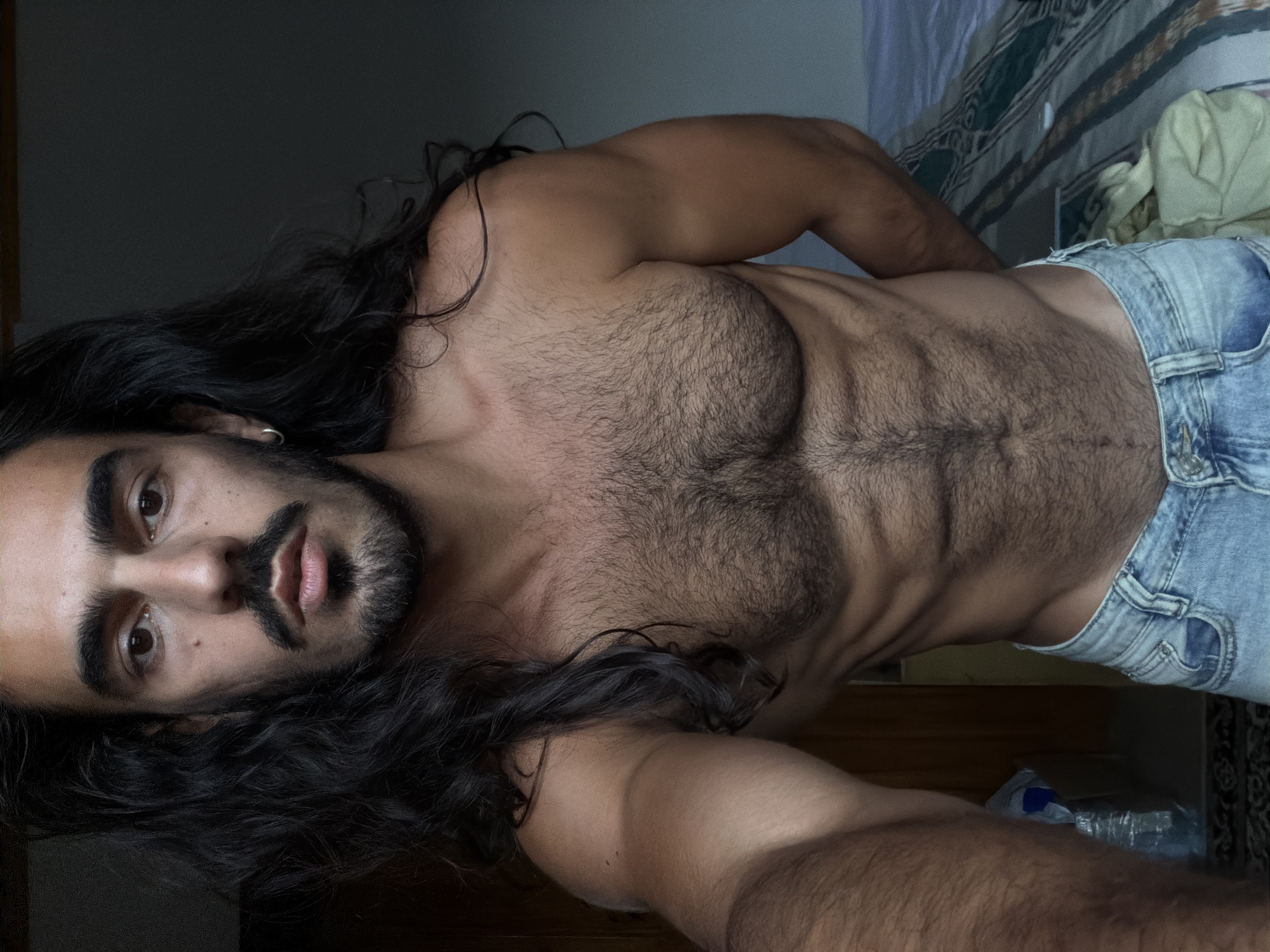 This vertical, waist-up portrait depicts a person, likely male, lying down indoors in North Kuta, Indonesia, and gazing directly into the camera. The subject has long, dark, wavy hair that frames his head and shoulders, complemented by a full beard and mustache. A small silver hoop earring is visible in his left ear. He displays a lean, muscular physique with prominent chest and abdominal hair that accentuates his defined abs and obliques. He is wearing light-wash denim shorts or jeans, worn low on his hips. The background is dimly lit and largely indistinct, showing hints of a patterned fabric, possibly a bedsheet or blanket, and a light-colored wall. Soft, directional lighting emphasizes his features and muscle definition. His expression is neutral with a direct, intense gaze. No text is visible in the image. A partially obscured plastic bottle is faintly visible in the lower right background.
